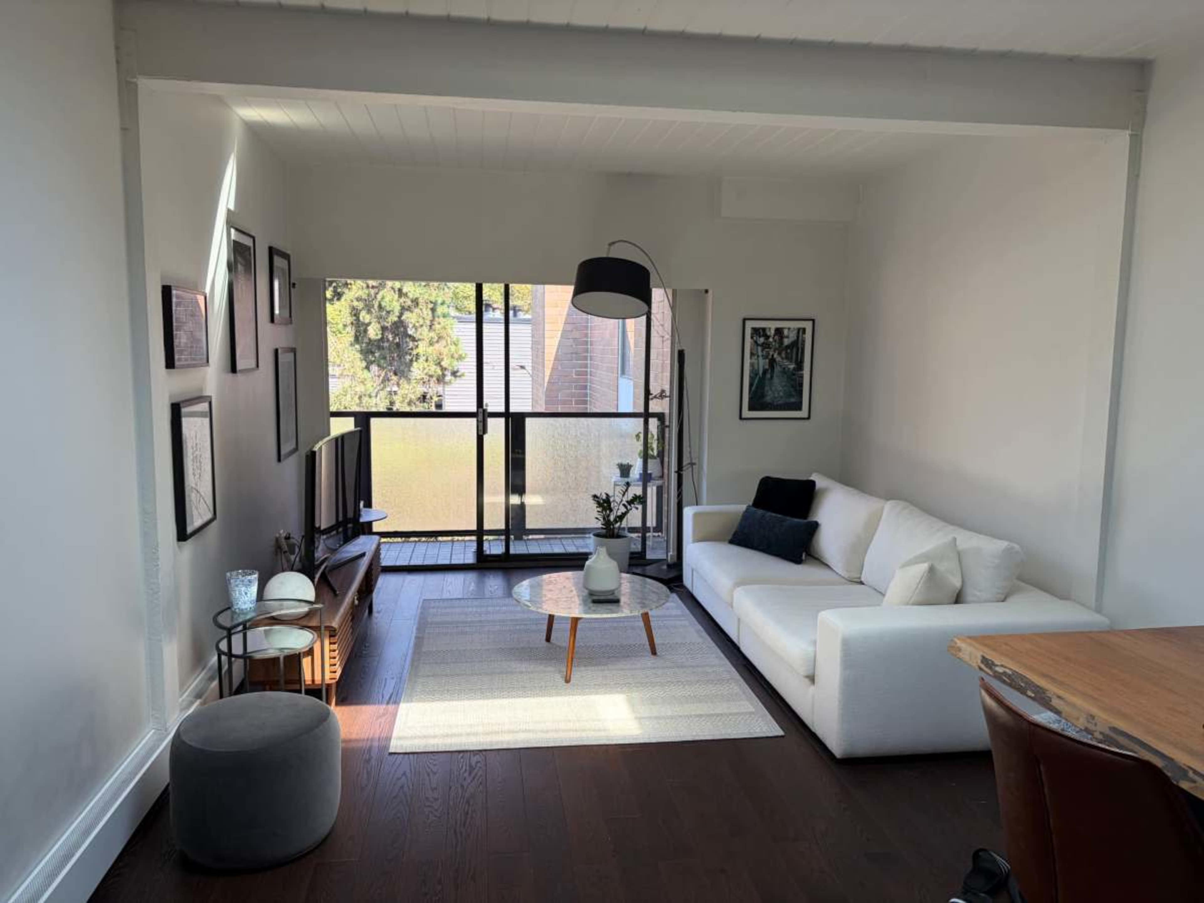 The image shows a modern, minimalist living room with a white sofa, a round coffee table, and a large window that allows natural light to flow in.