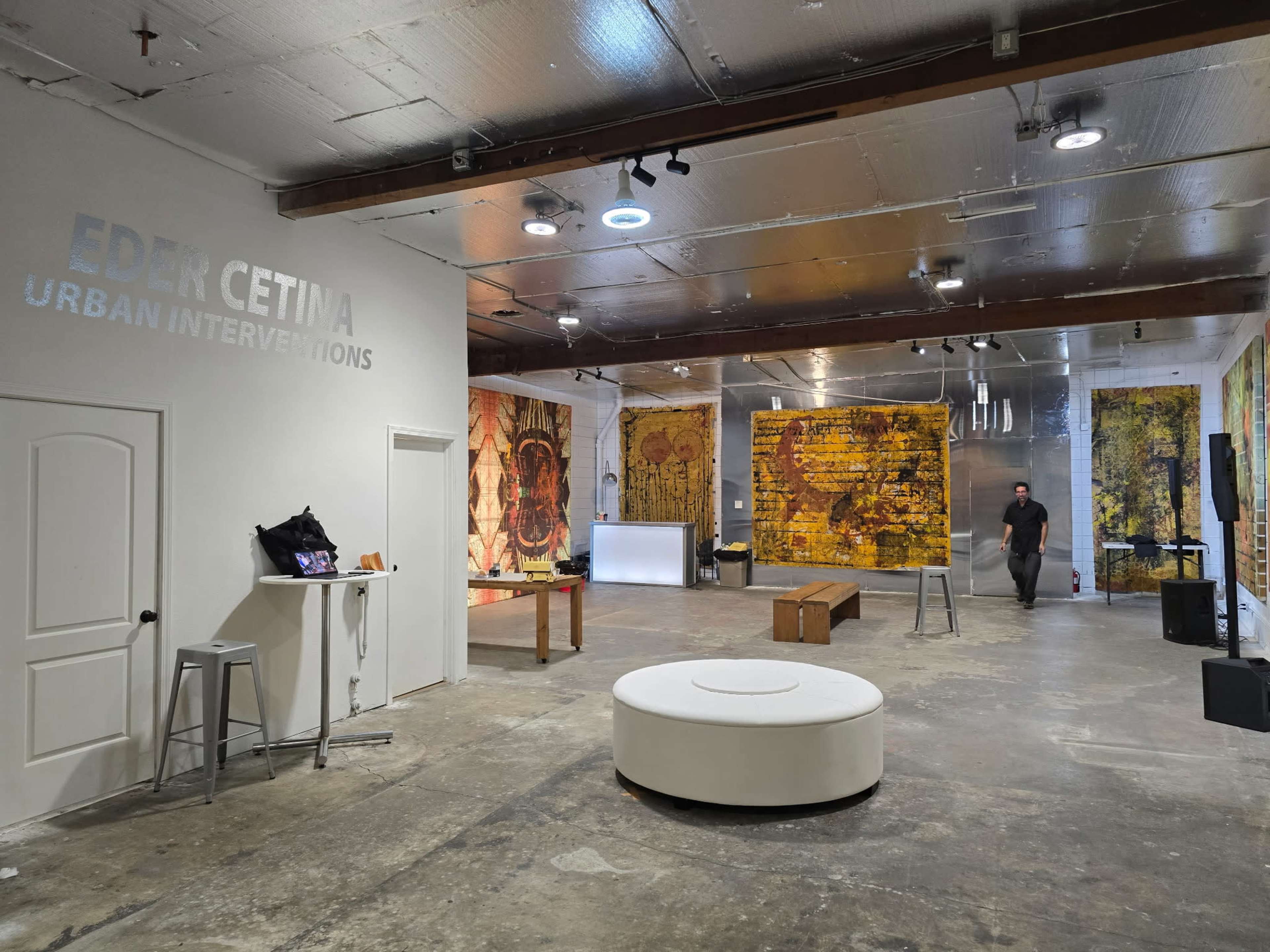 The image shows a spacious art gallery featuring large, colorful murals on the walls, a white circular seating area in the center, and various furniture pieces arranged throughout the space.