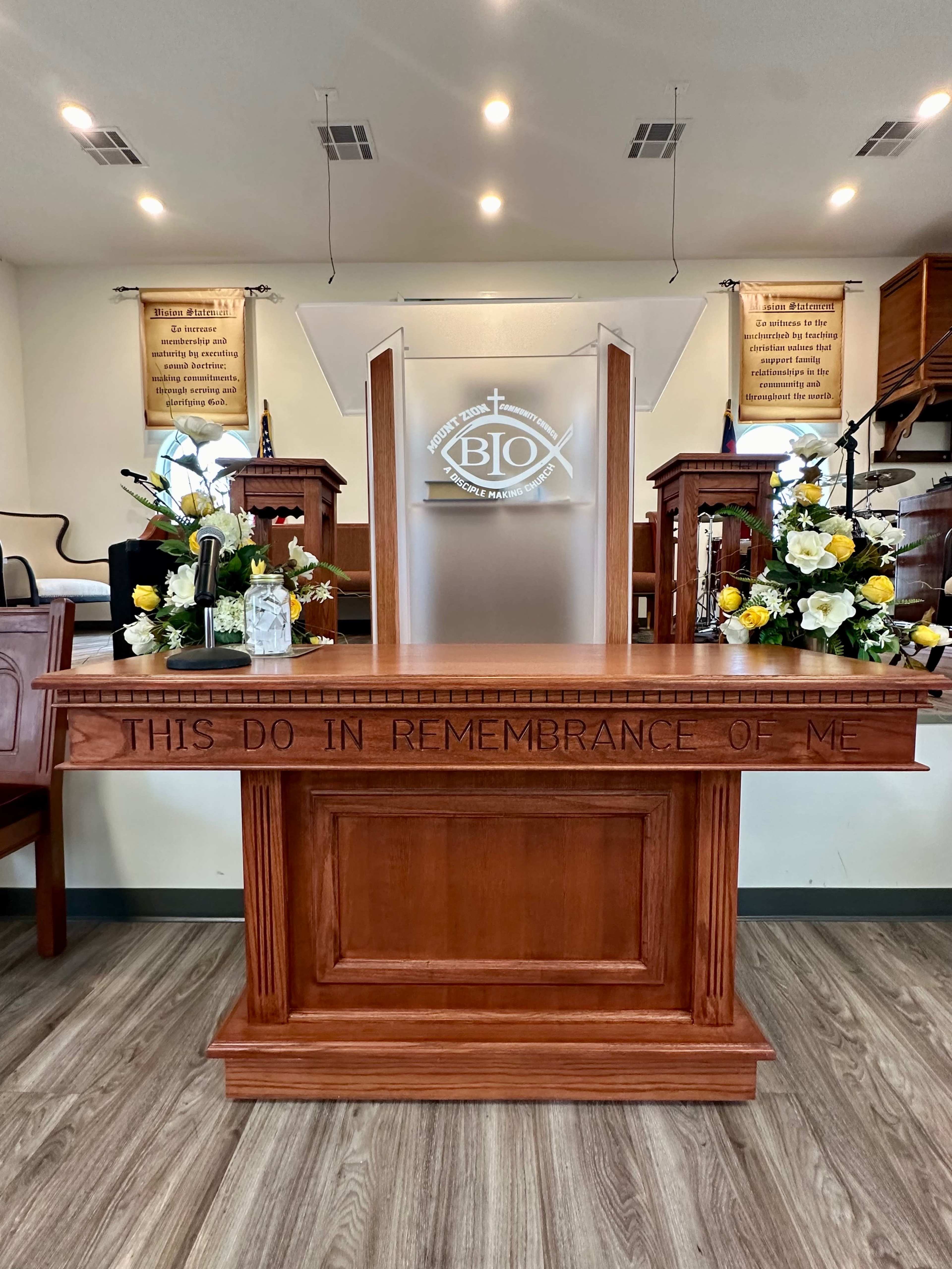 The image shows a wooden altar with the inscription "THIS DO IN REMEMBRANCE OF ME," adorned with floral arrangements, set in a church interior.