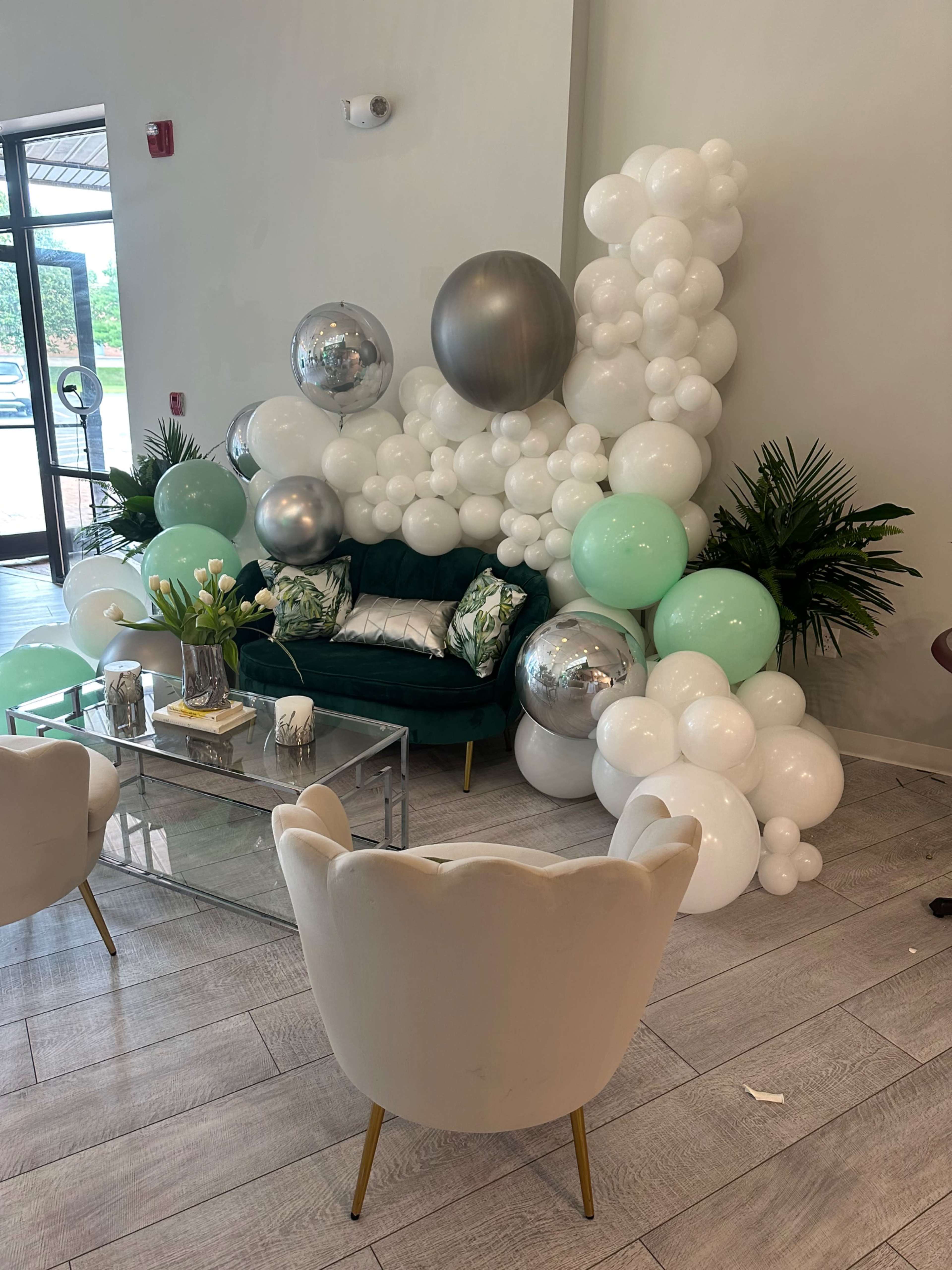 A cozy seating area with a green couch surrounded by a decorative arrangement of white and mint balloons, silver spheres, and potted plants.