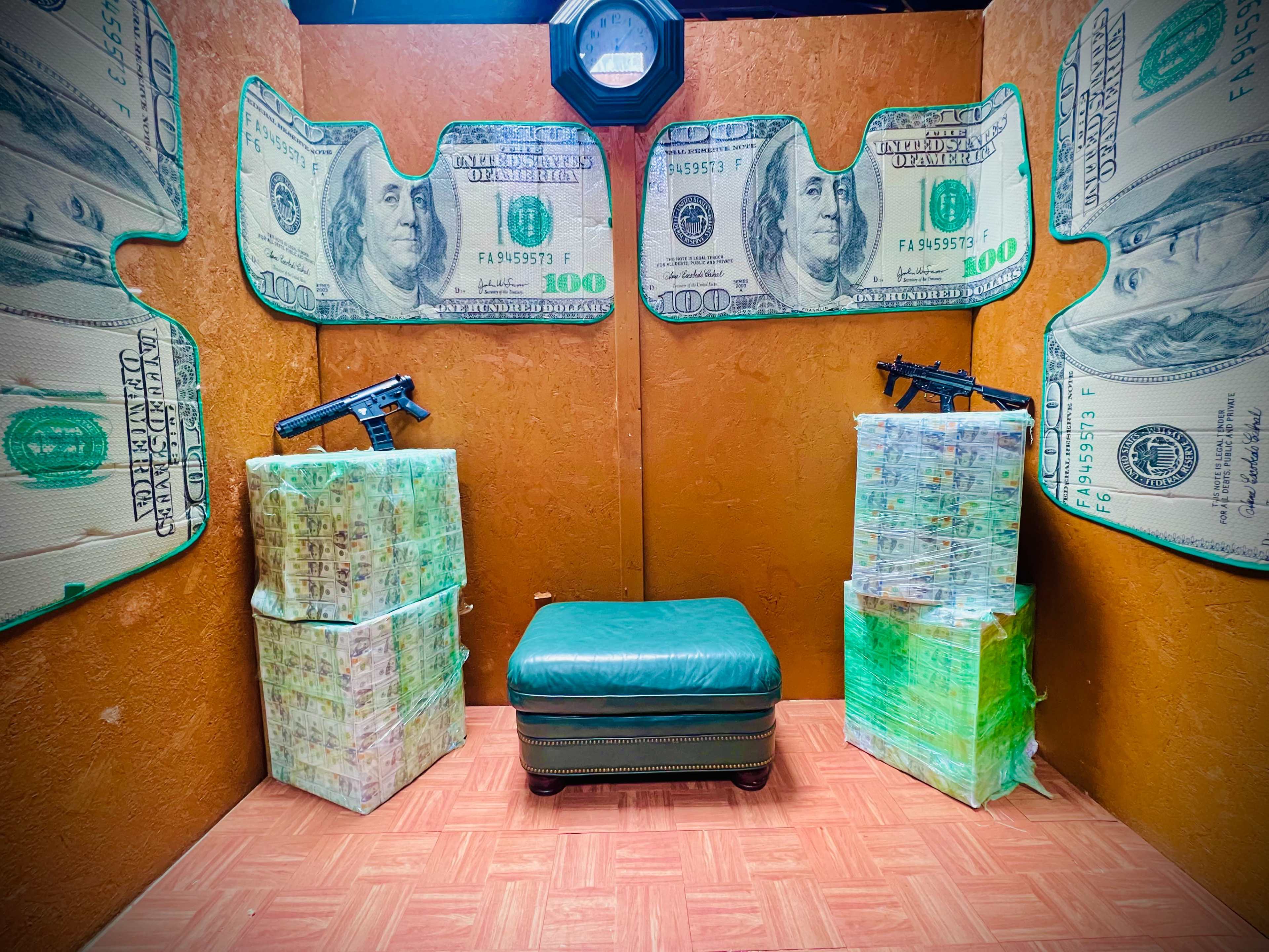 The image shows a small room decorated with large dollar bill motifs, featuring stacks of money on either side of a green ottoman, with two handguns displayed on the walls.