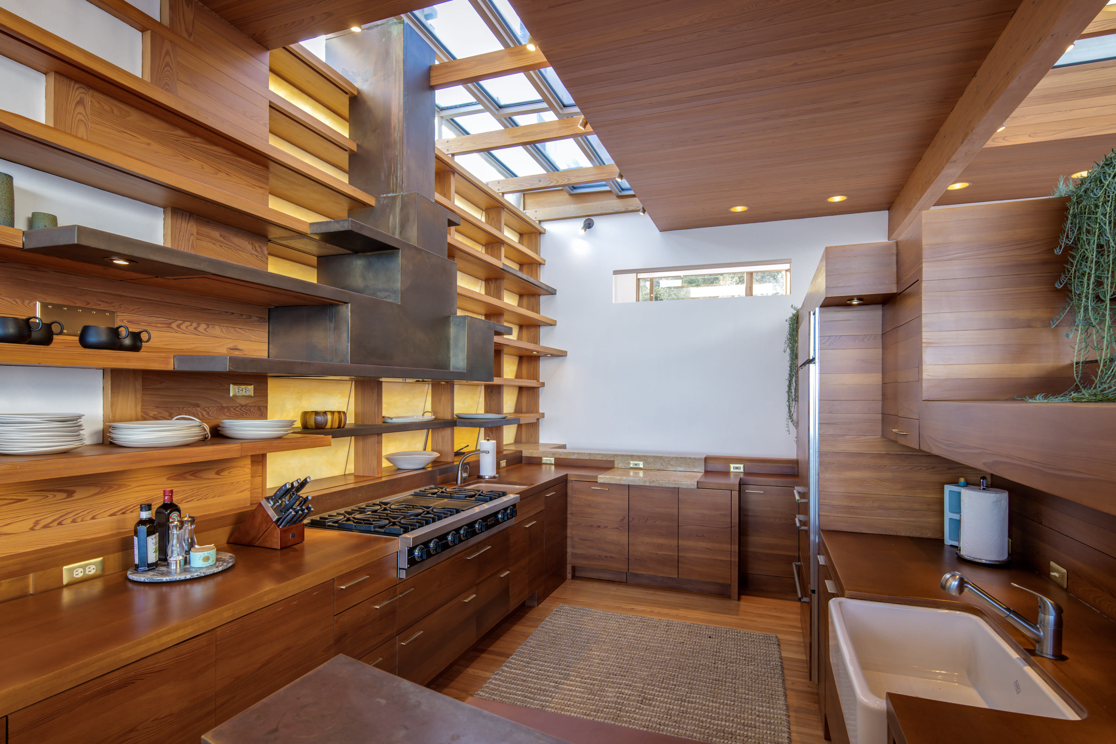 A modern kitchen with wooden cabinetry, open shelving, a stainless steel stove, and a sink under a skylight.