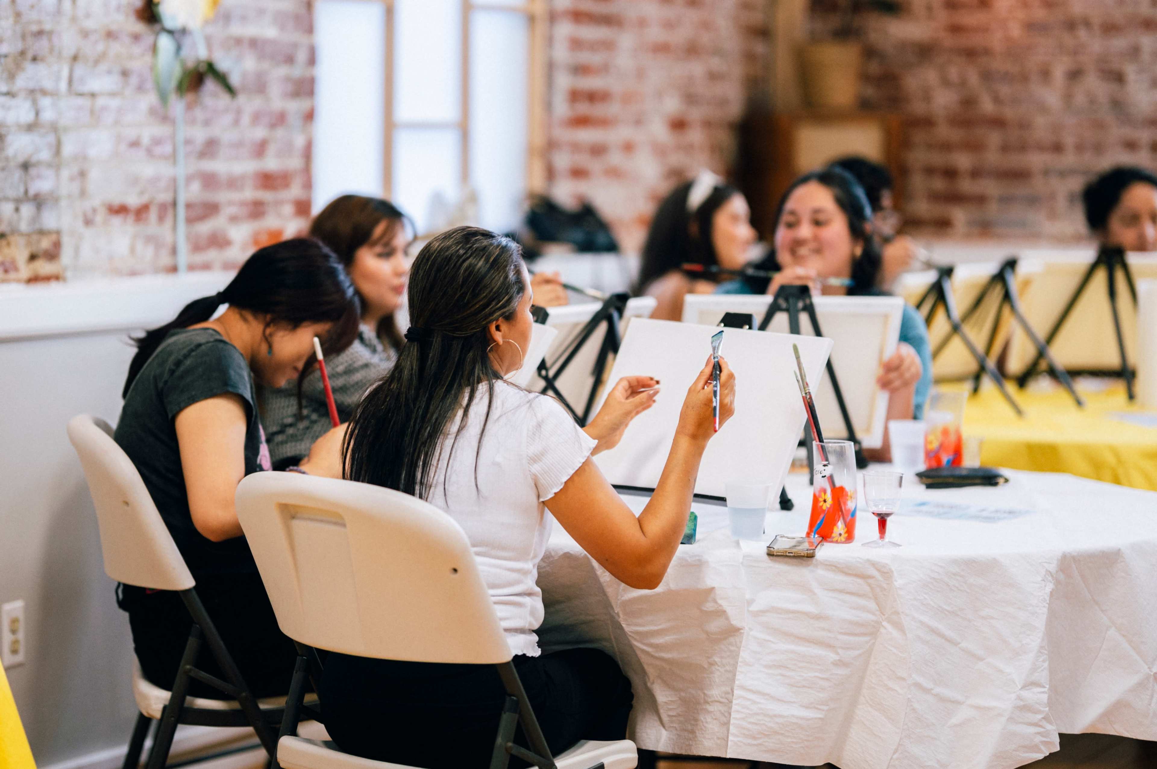 A group of people sits at tables, engaged in a painting session with canvases and paint supplies.