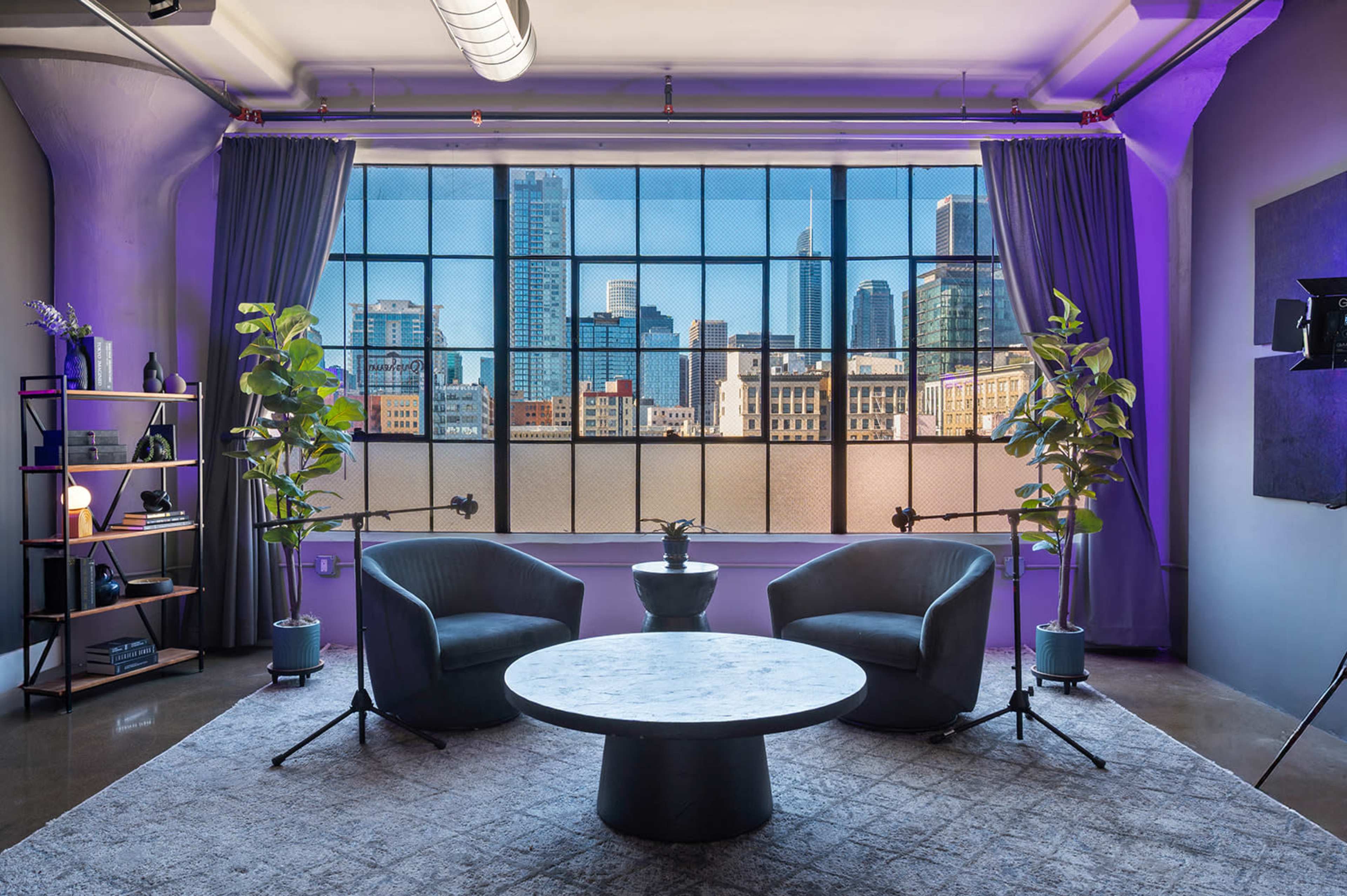 The image shows a modern interior with two chairs facing a round table, framed by large windows overlooking a city skyline.