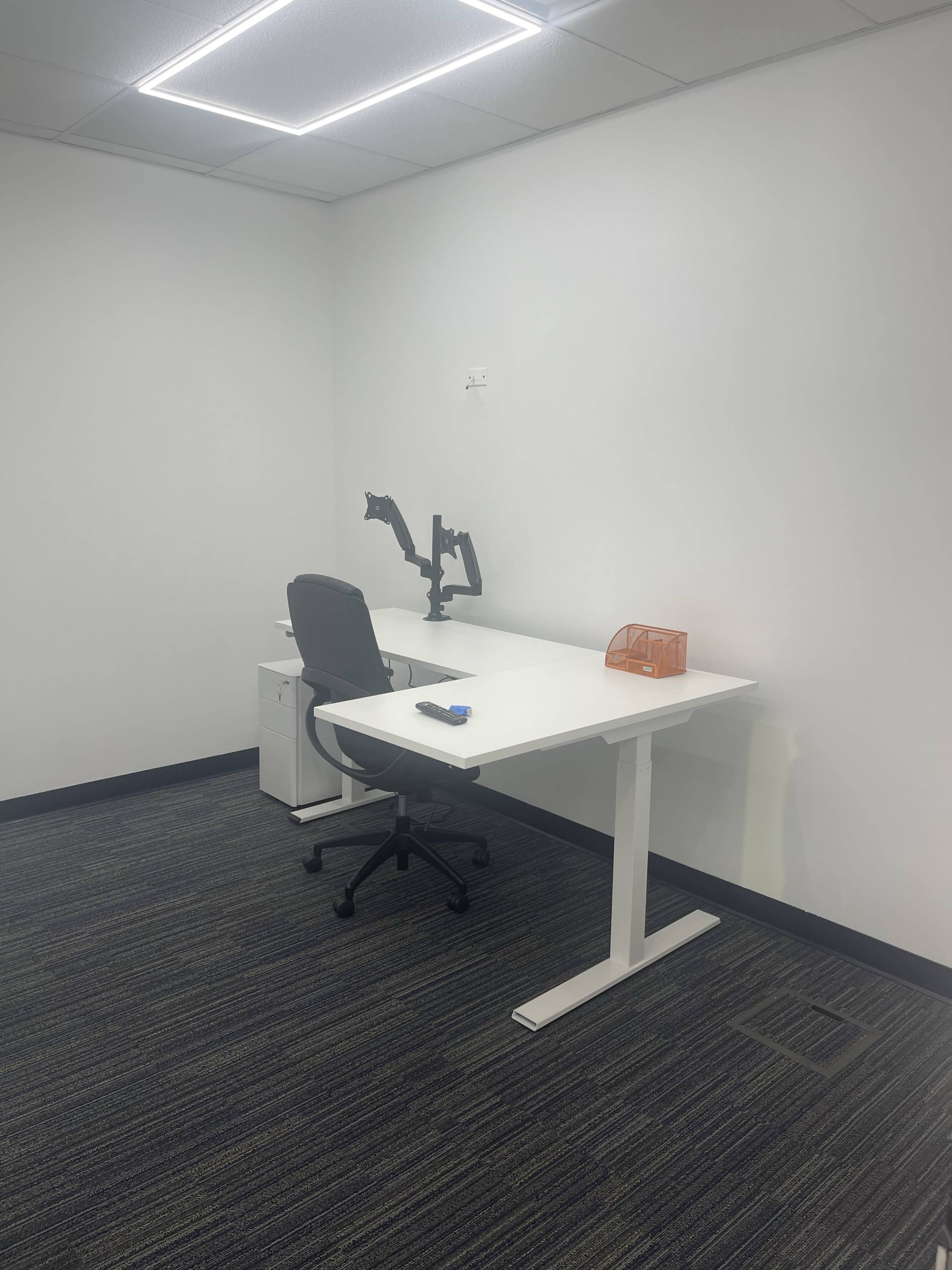 A minimalist office space featuring a white adjustable desk, a gray office chair, a monitor mount, and a small orange storage container against a plain white wall.