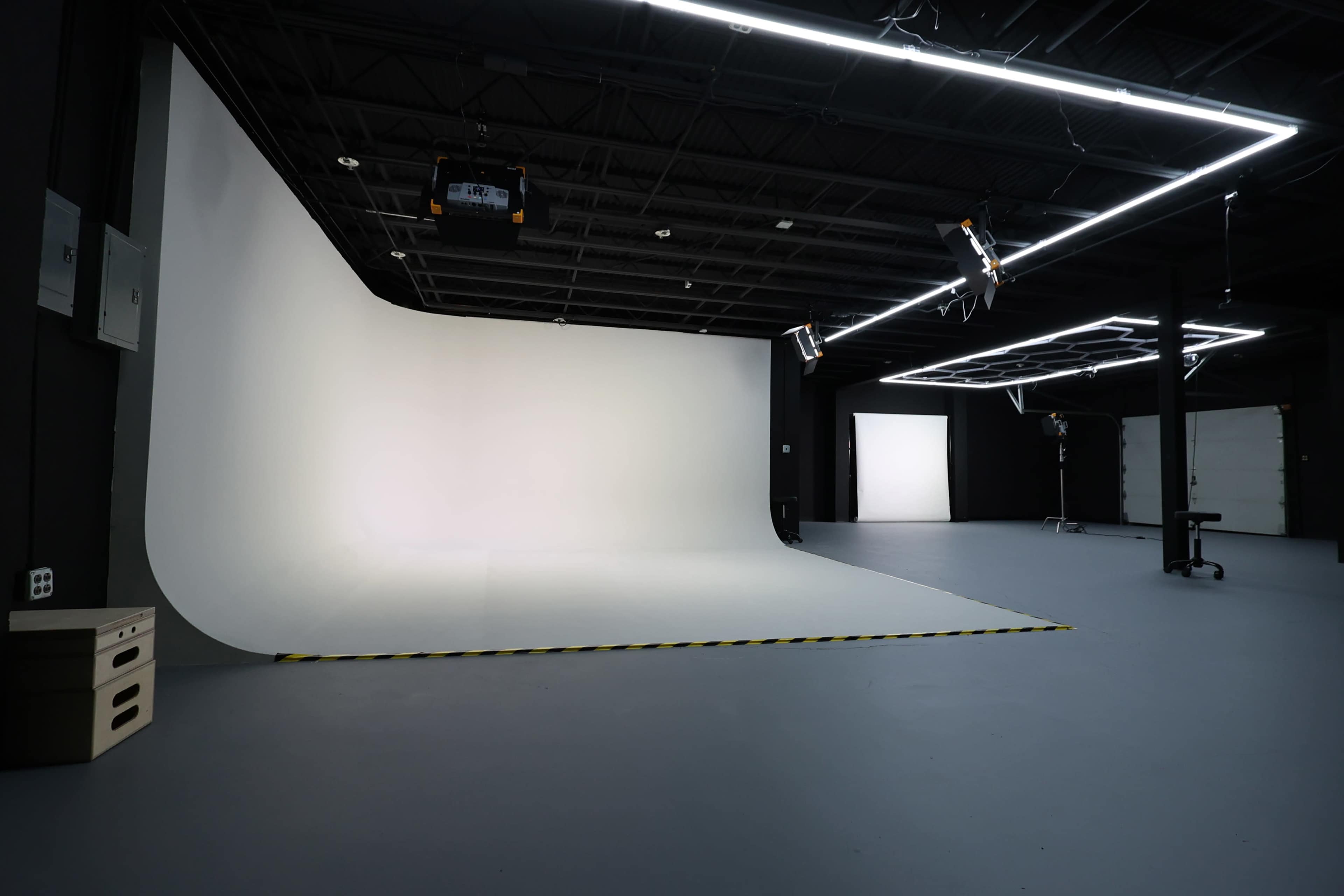 The image shows a photography studio with a white seamless backdrop, overhead lighting, and a clean, uncluttered floor space.