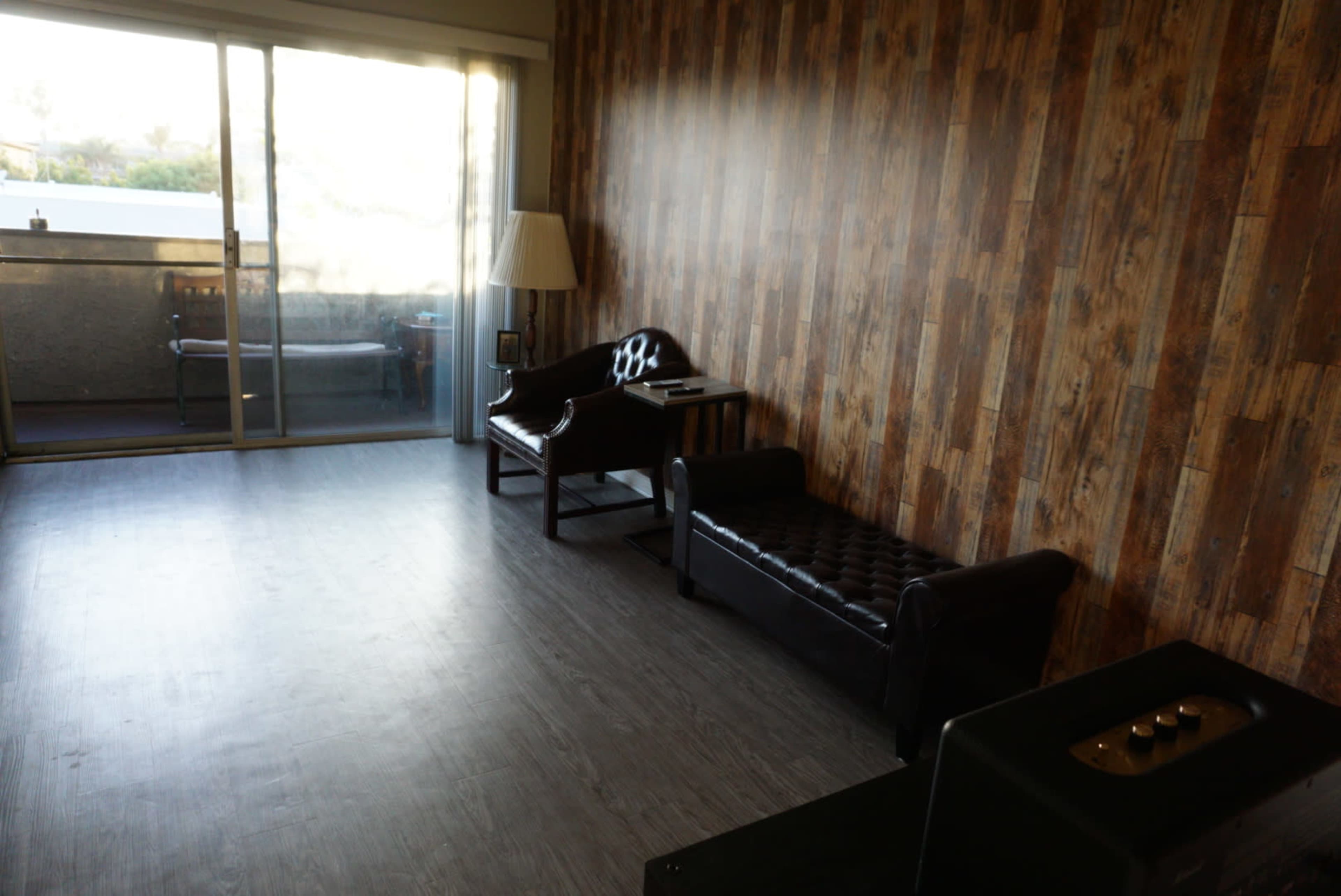 A minimalist living room with a wooden-paneled wall, a leather chaise lounge, a chair, and a lamp near a large sliding glass door that leads to a balcony.