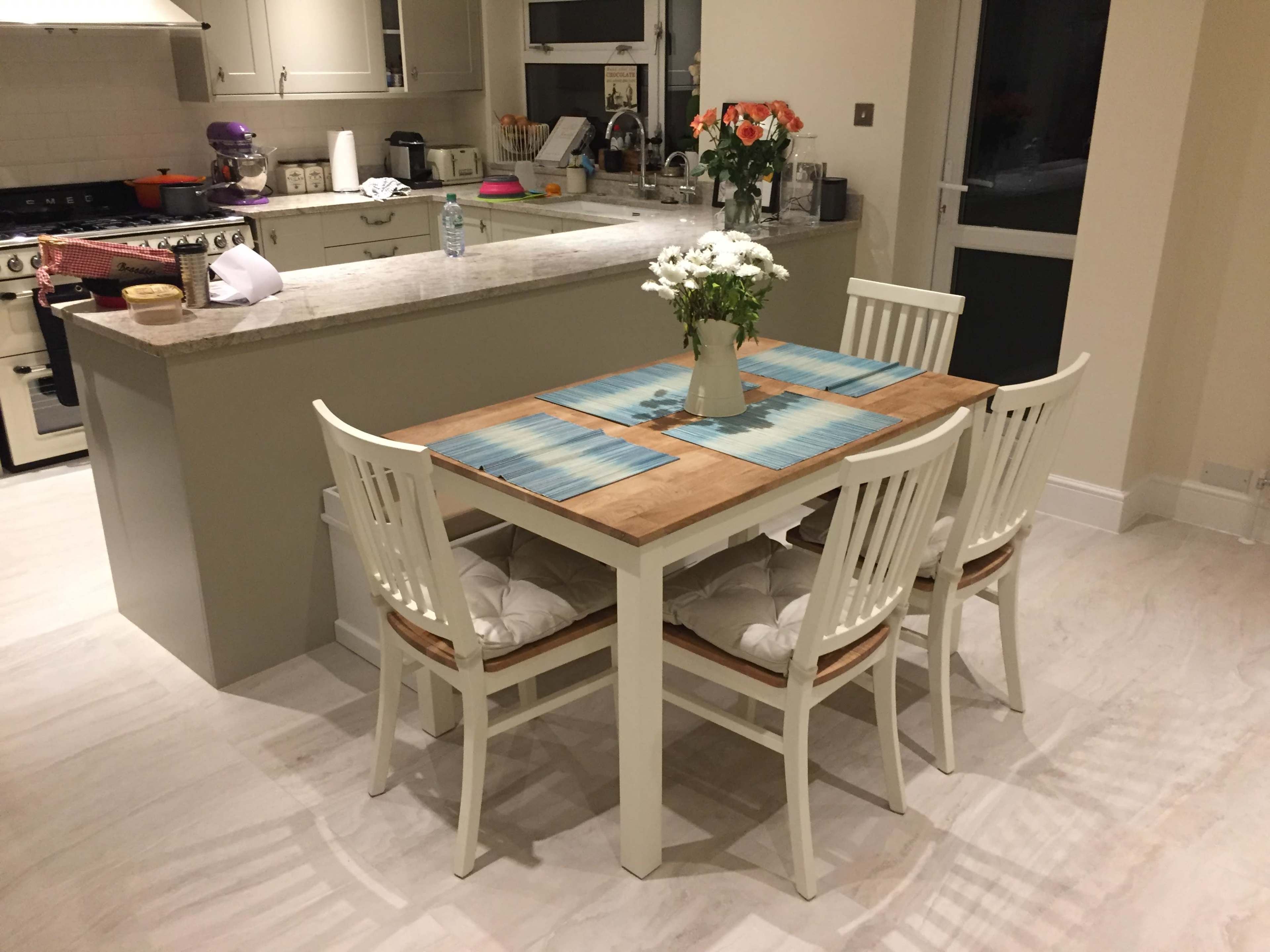 A dining table with four chairs is set in a kitchen, featuring a centerpiece of flowers and placemats, while an island with a sink and various kitchen items is visible in the background.