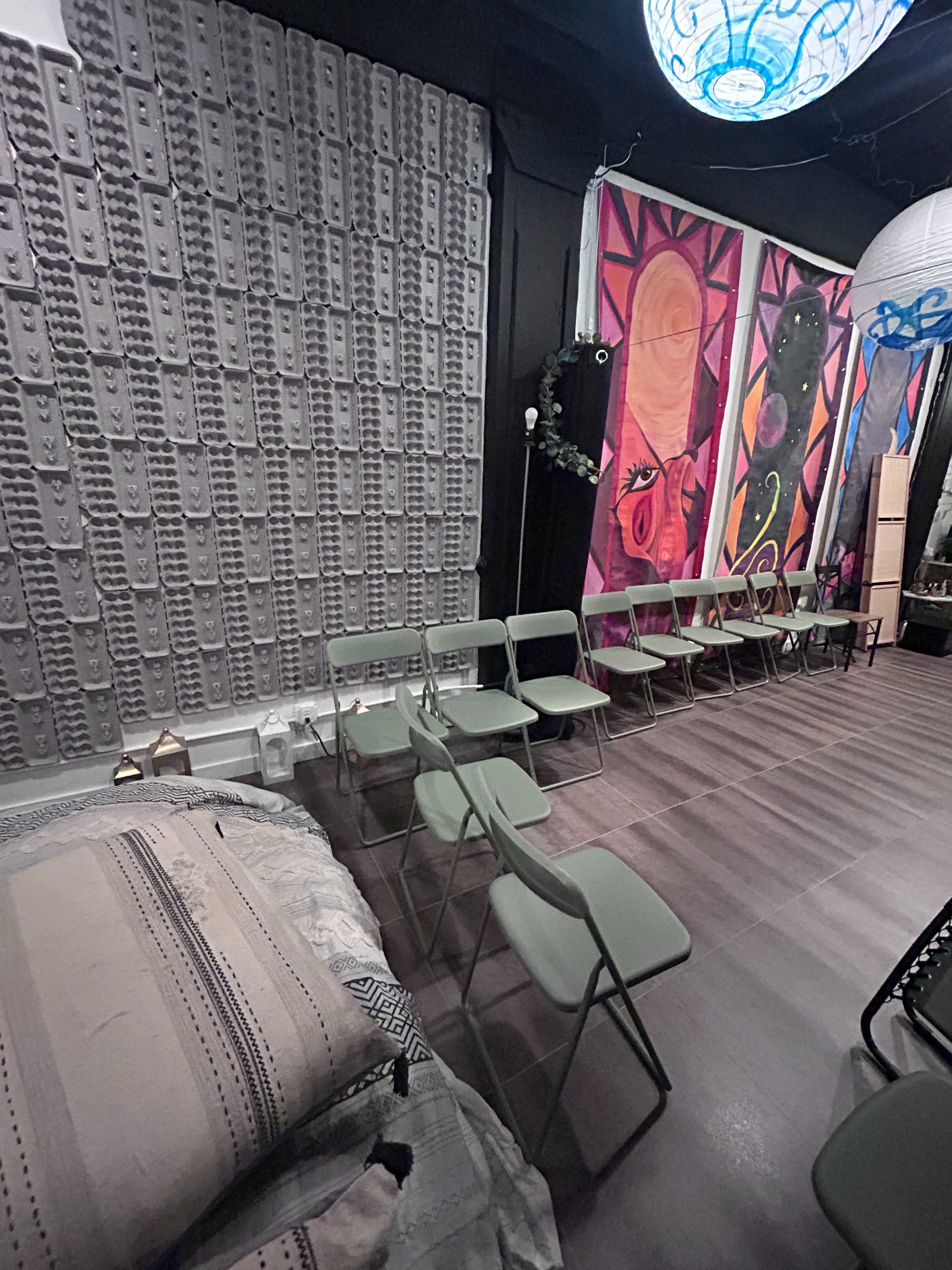 The image shows a room with several folding chairs arranged in a row, a decorative wall with a textured design, and colorful murals depicting abstract figures.