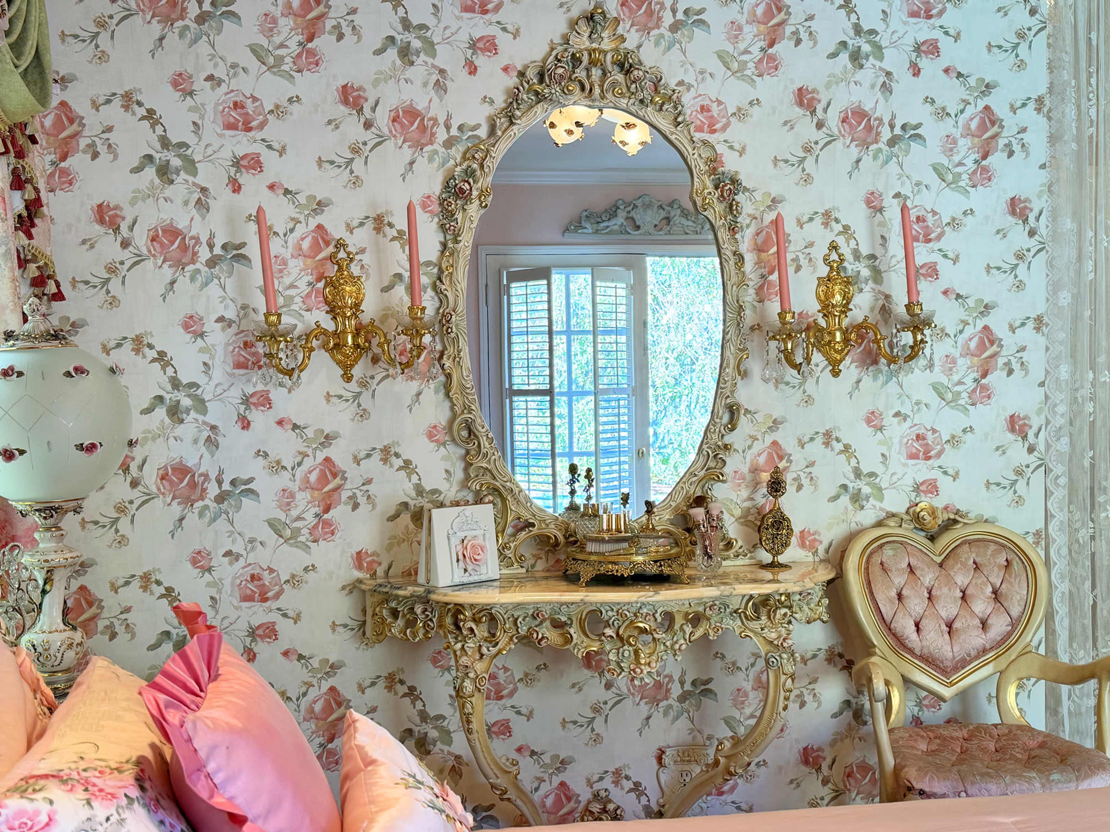 The image shows a decorative bedroom featuring a floral wallpaper, an ornate mirror, a console table with decorative items, and a heart-shaped chair.
