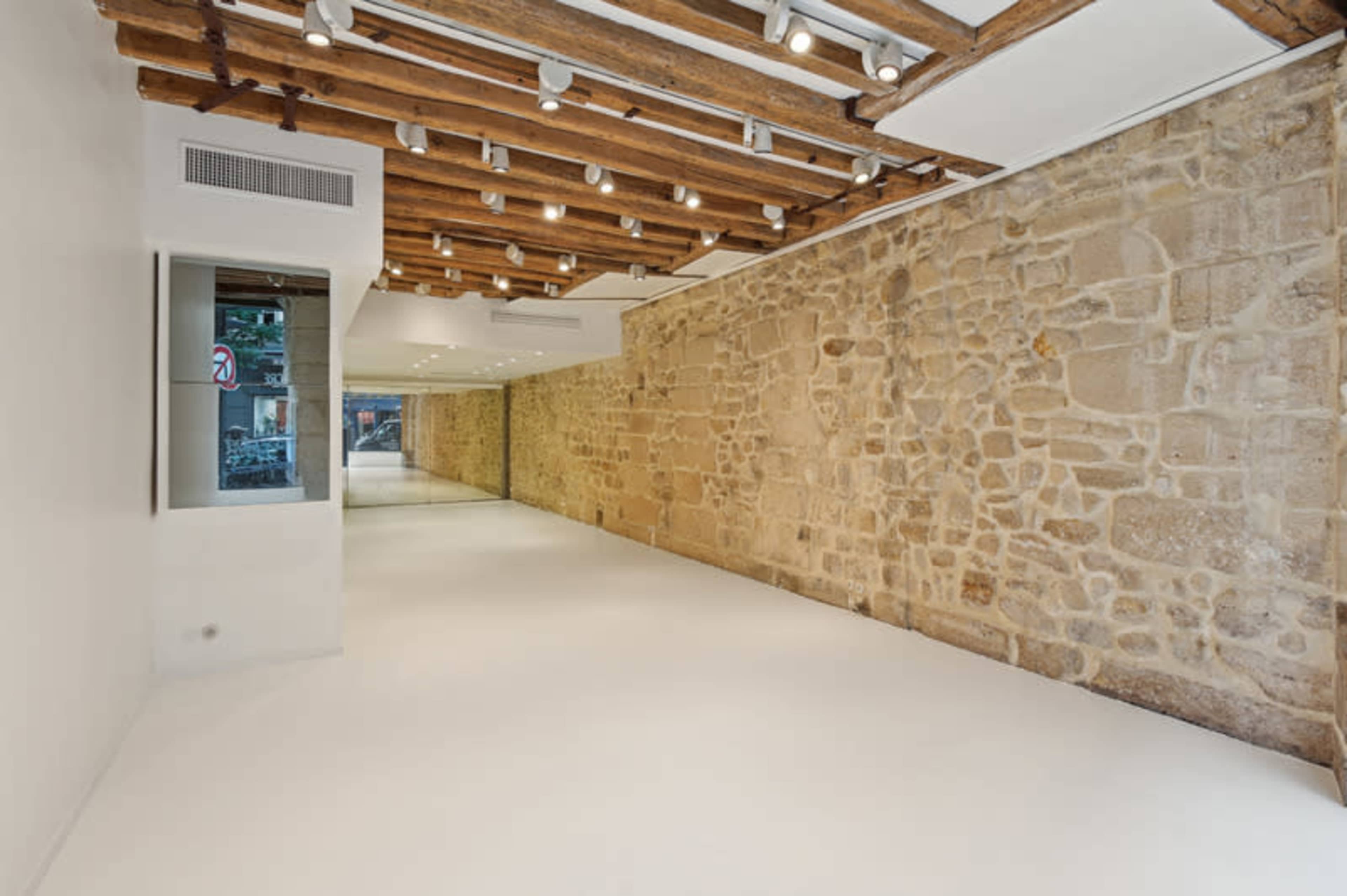 The image shows a spacious interior room featuring a combination of exposed wooden beams and a stone wall, with white floors and bright lighting.