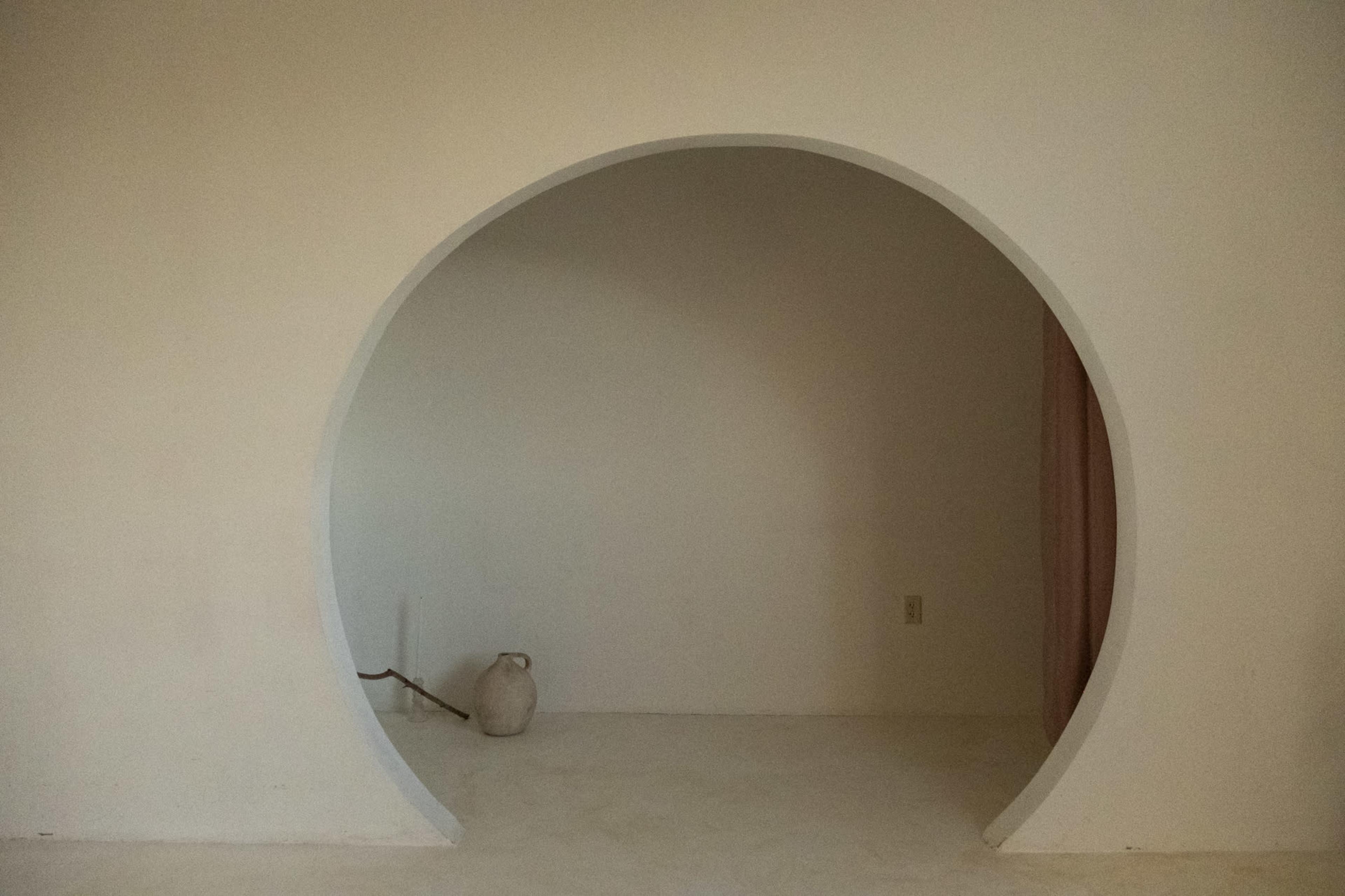 The image shows a circular archway in a minimalist room, leading to a small, empty space with a ceramic jug placed on the floor.