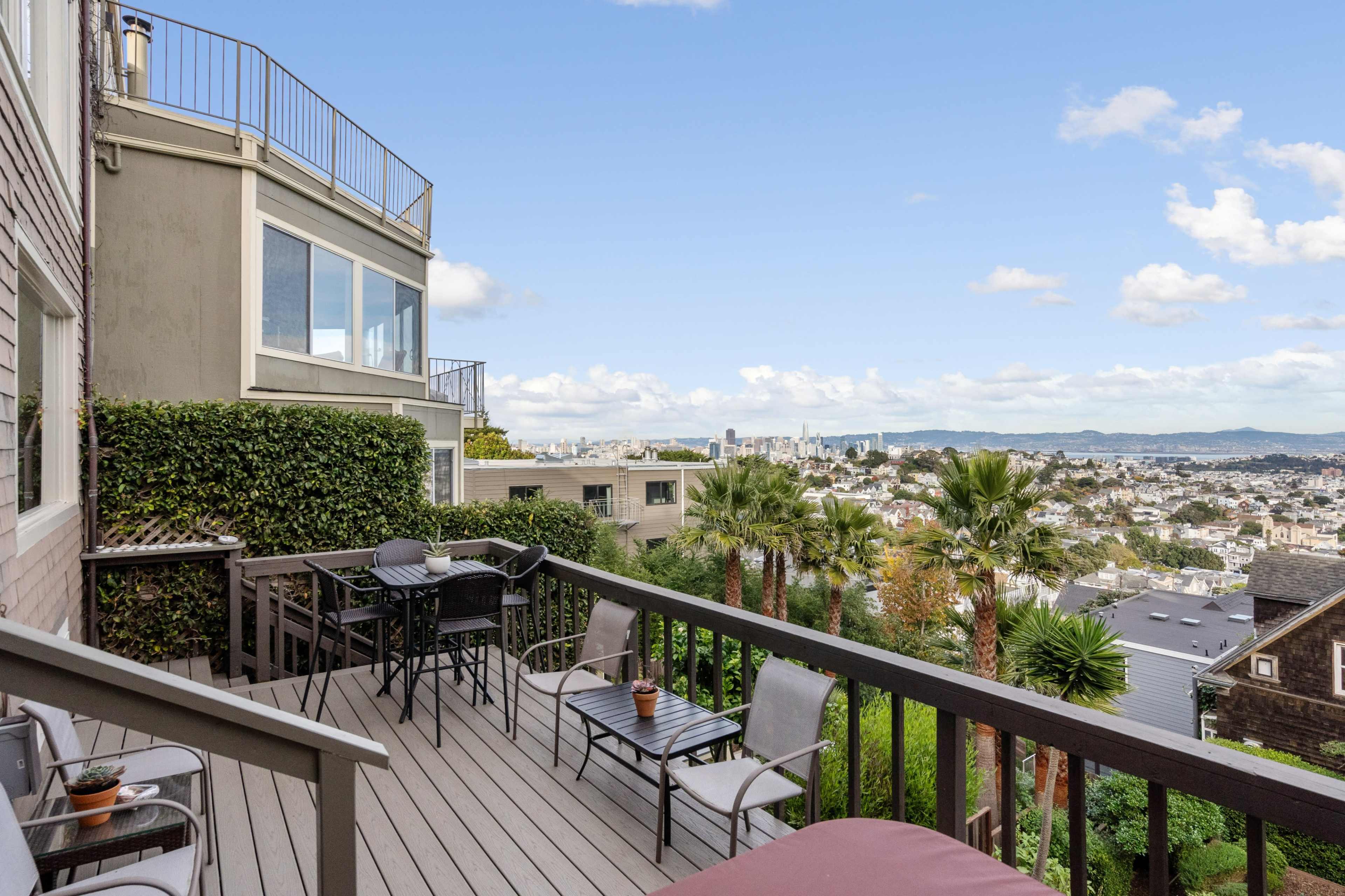 The image shows a wooden deck with seating that overlooks a cityscape, featuring palm trees and distant hills under a partly cloudy sky.