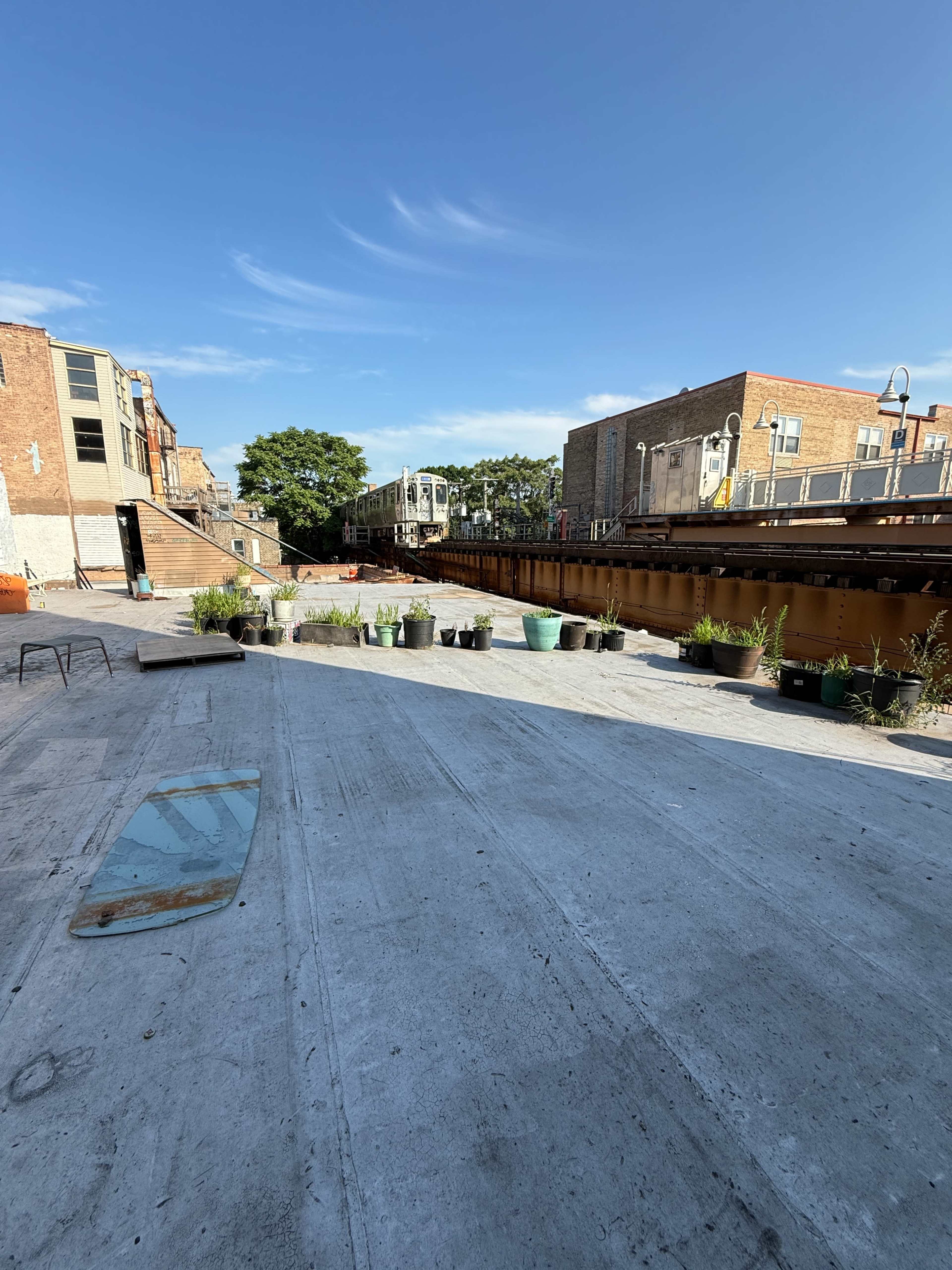 The scene shows a concrete pathway bordered by potted plants, leading to a canal with a train track overhead and buildings lining the sides.