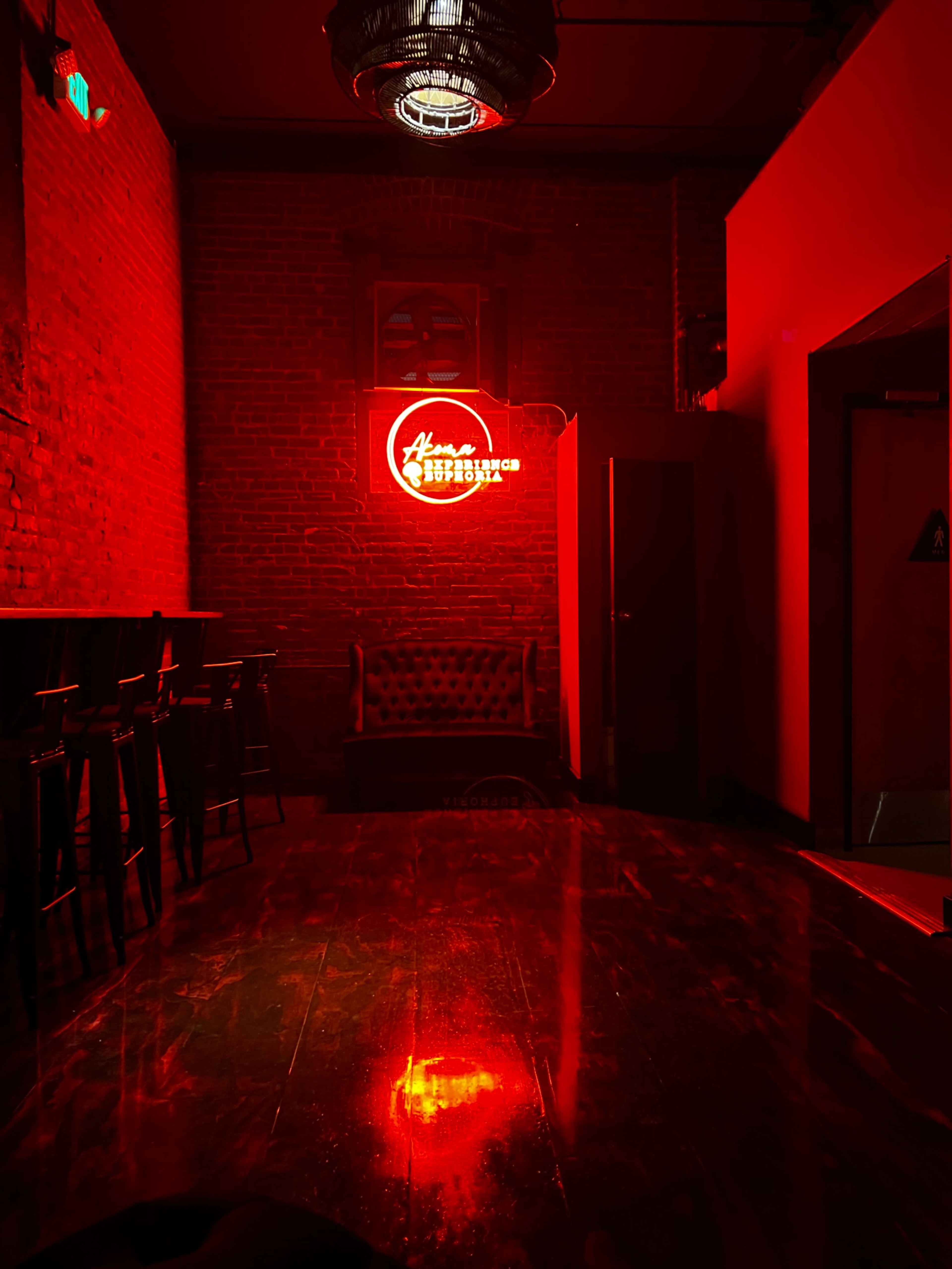 A dimly lit room featuring a red neon sign, dark wooden furniture, and exposed brick walls.
