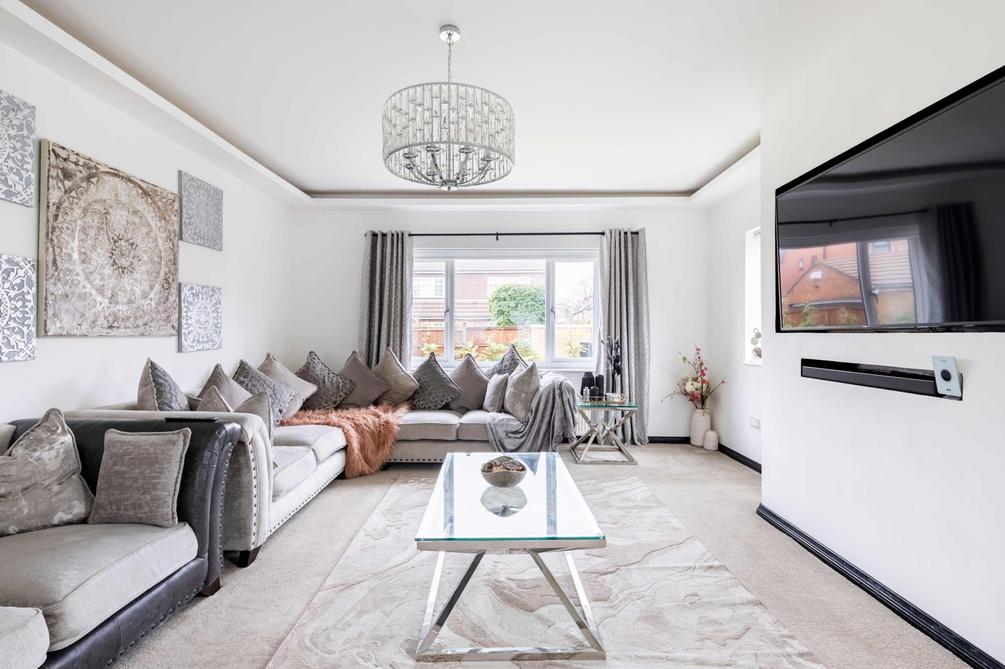 The living room features a gray sectional sofa with decorative pillows, a glass coffee table, and large windows allowing natural light to fill the space.