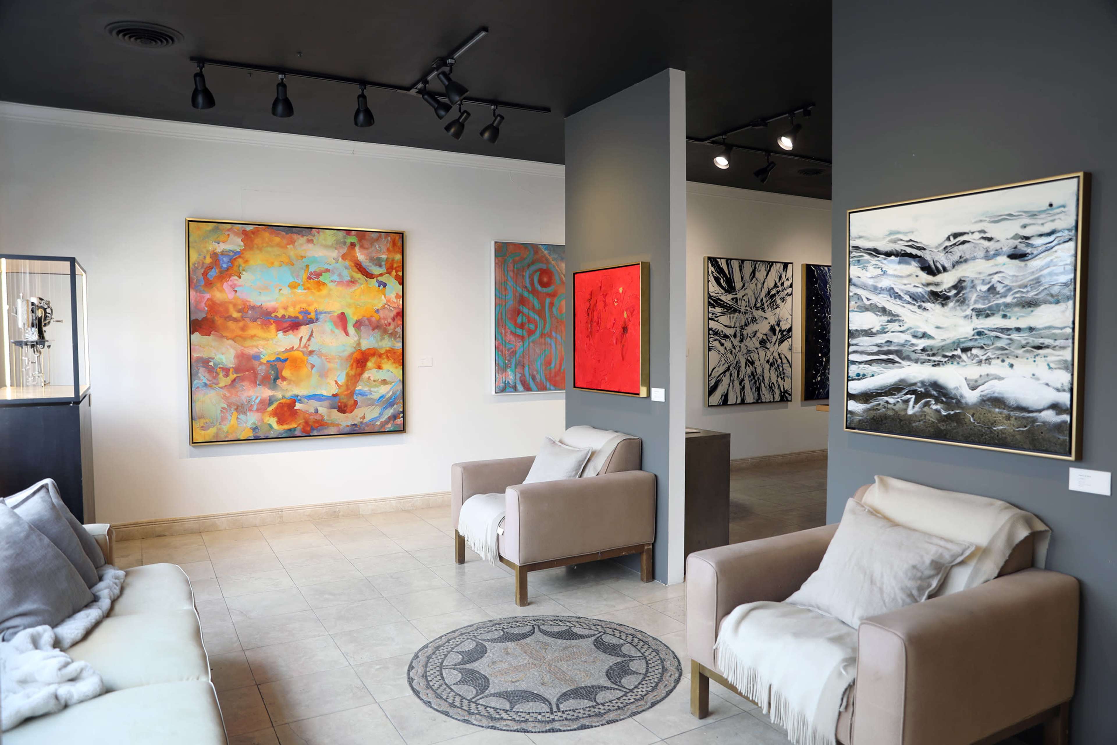 The image shows an art gallery featuring abstract paintings on the walls, with two chairs and a decorative rug in the foreground.