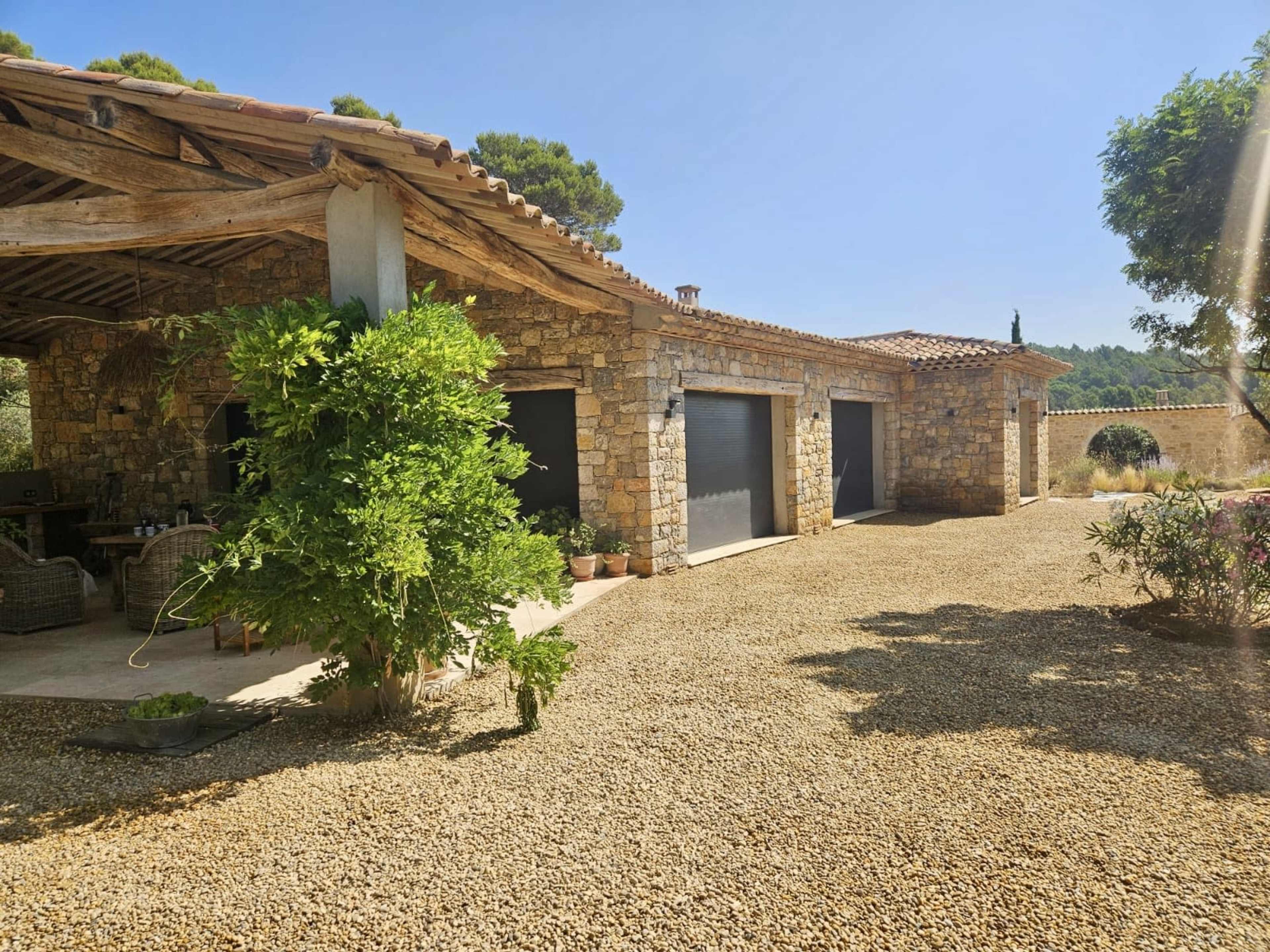 A stone house with a rustic roof and gravel driveway features three garage doors and a landscaped area with potted plants.