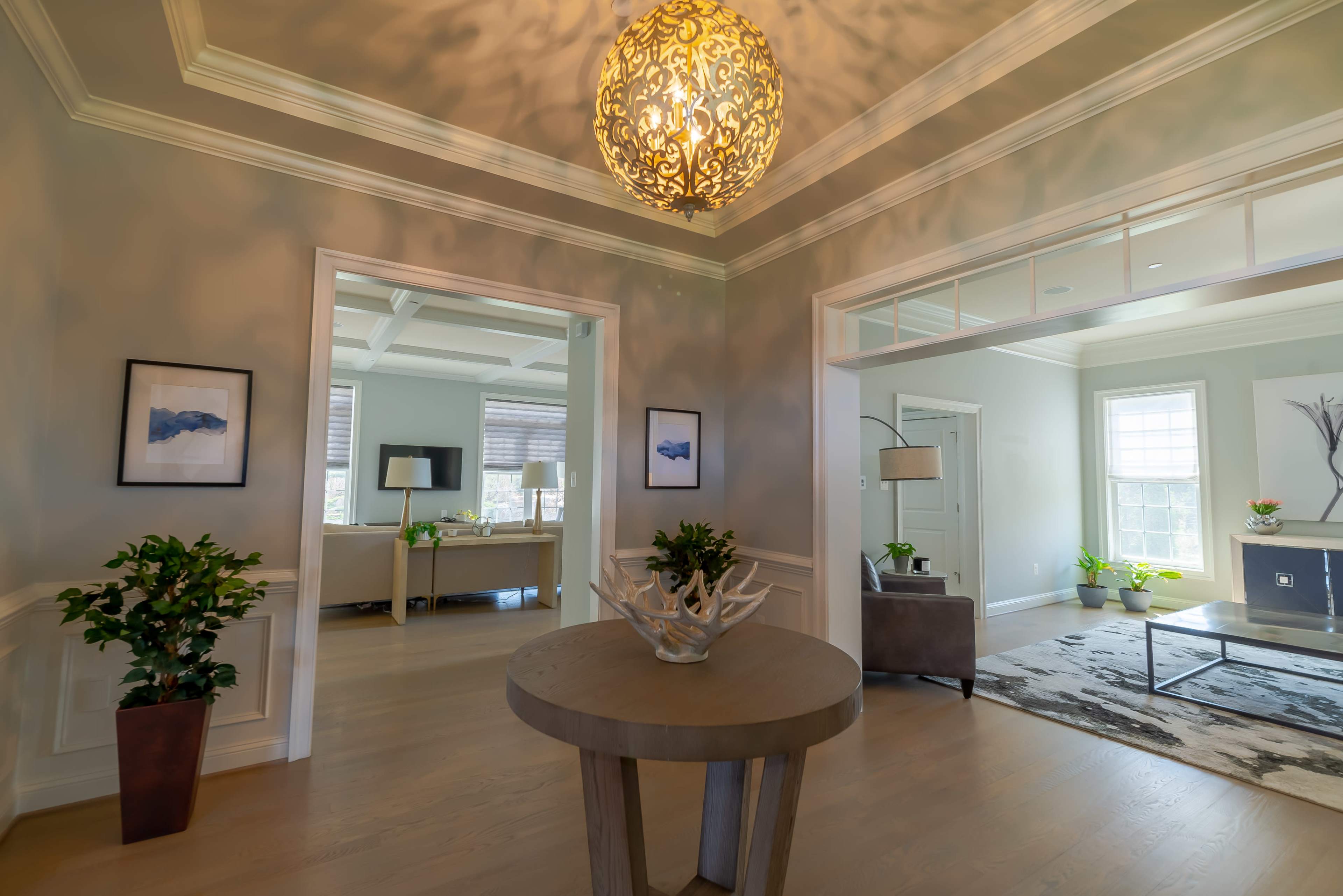 A spacious, well-lit foyer with a decorative light fixture, a round table, and potted plants, leading to an adjacent living area.