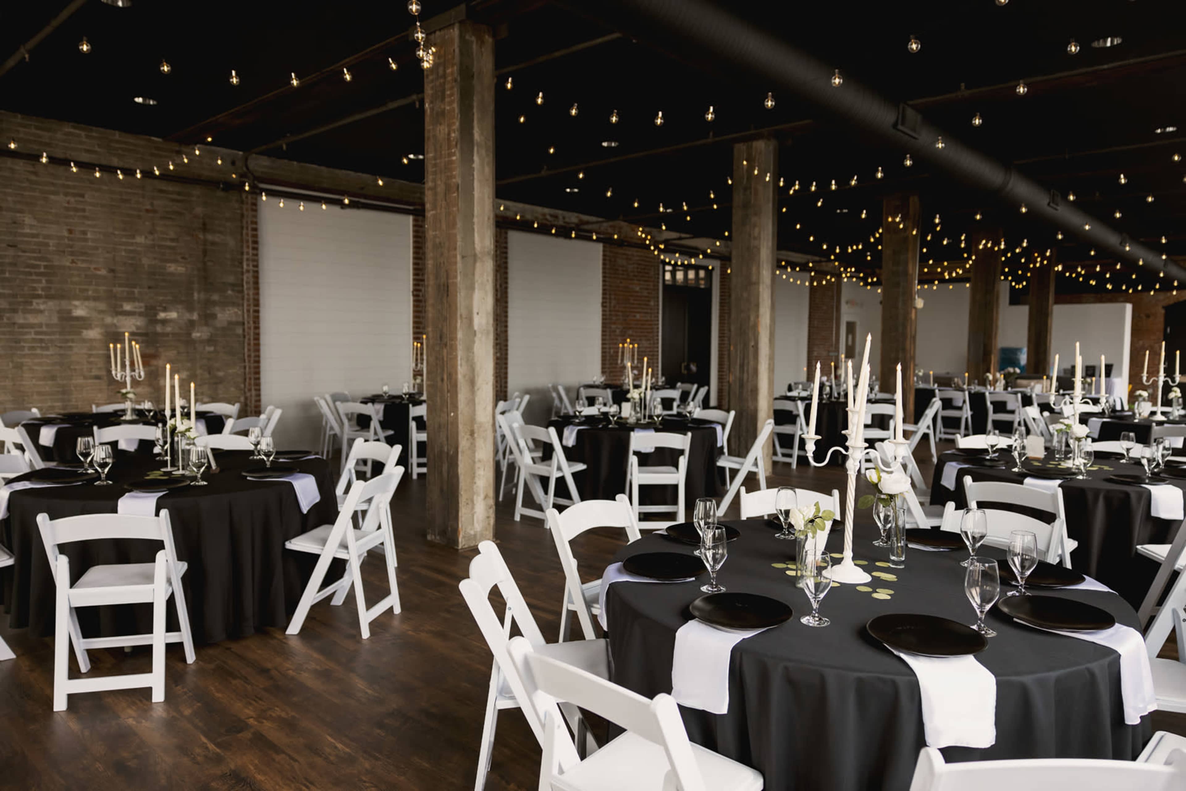 A decorated event space with round tables covered in black tablecloths, white chairs, and string lights overhead.