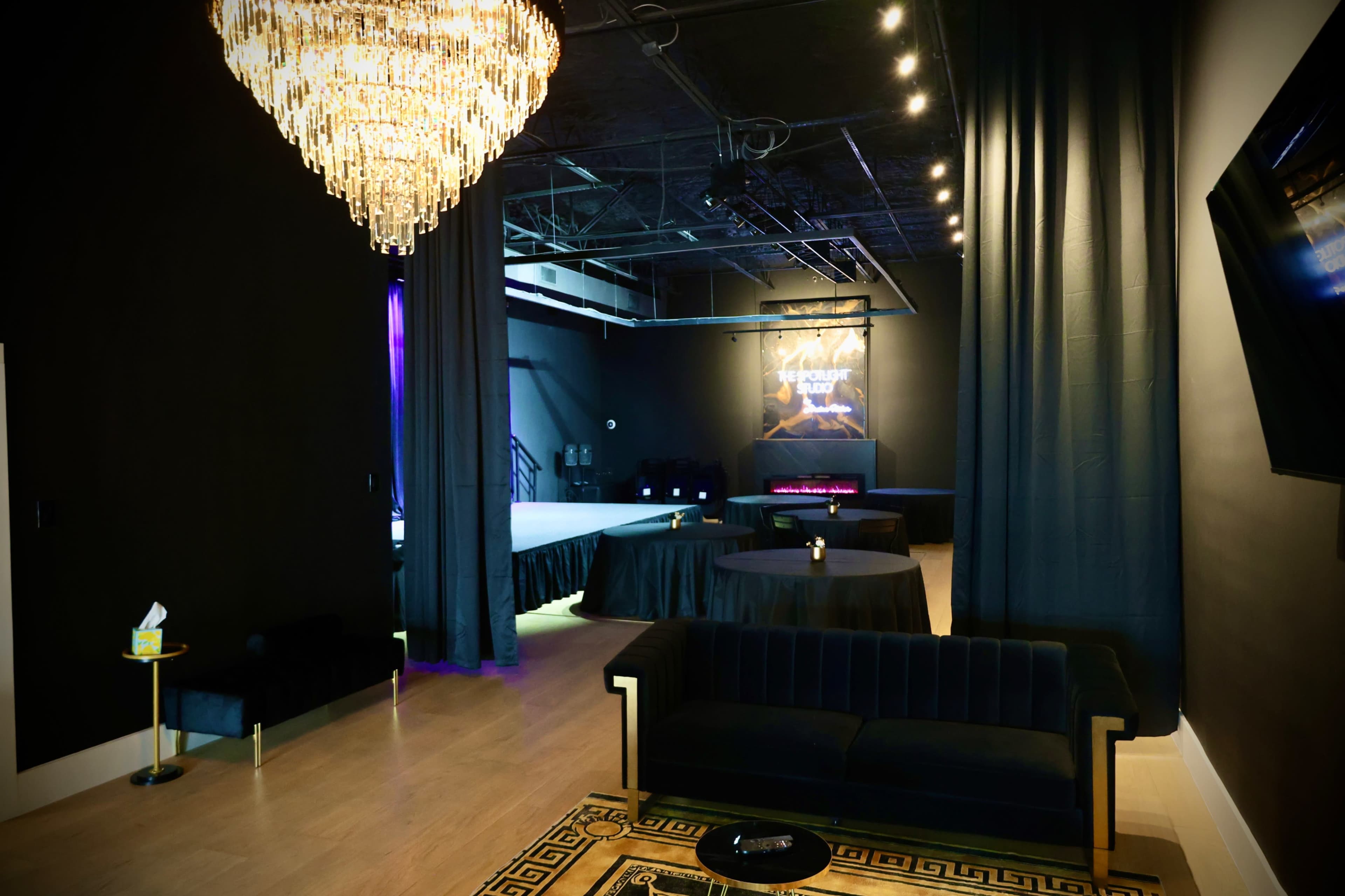 The image shows a modern event space with a stage in the background, surrounded by elegant seating and a large chandelier overhead.