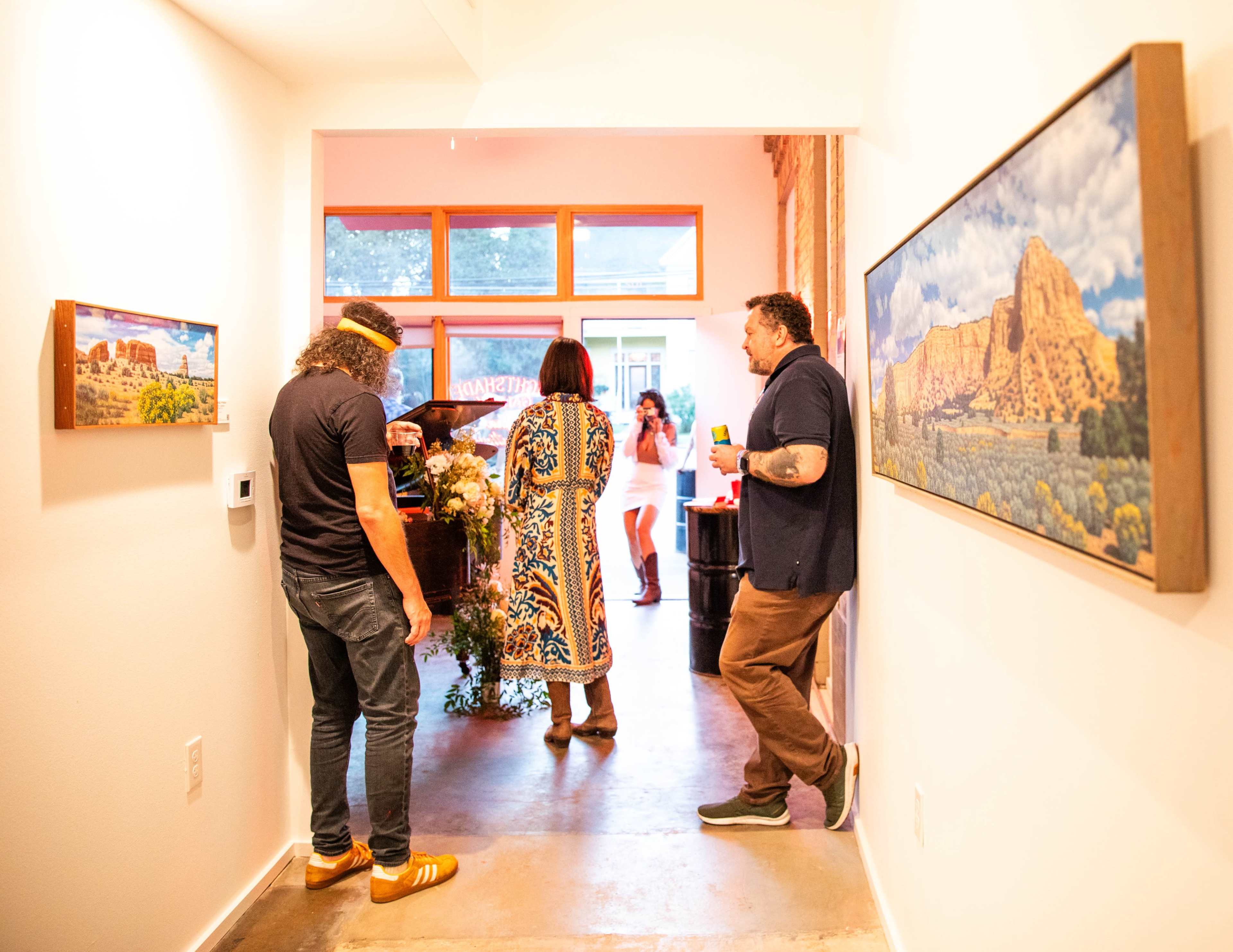Two people stand in a hallway, while another person enters a brightly lit room that features artwork on the walls and a gathering of people.