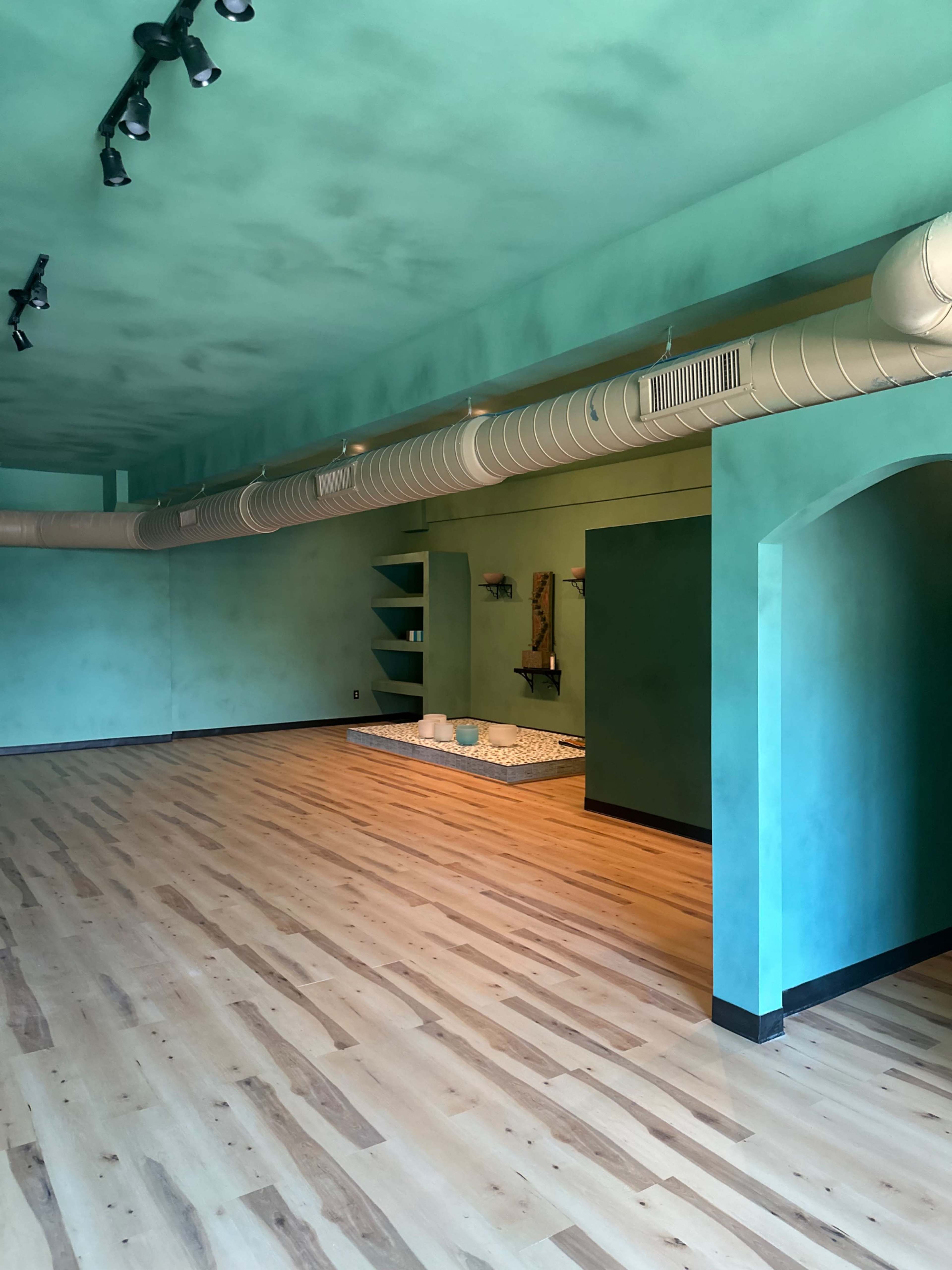 An empty room with teal walls, wooden flooring, and a central area featuring a decorative mat and a small water feature.