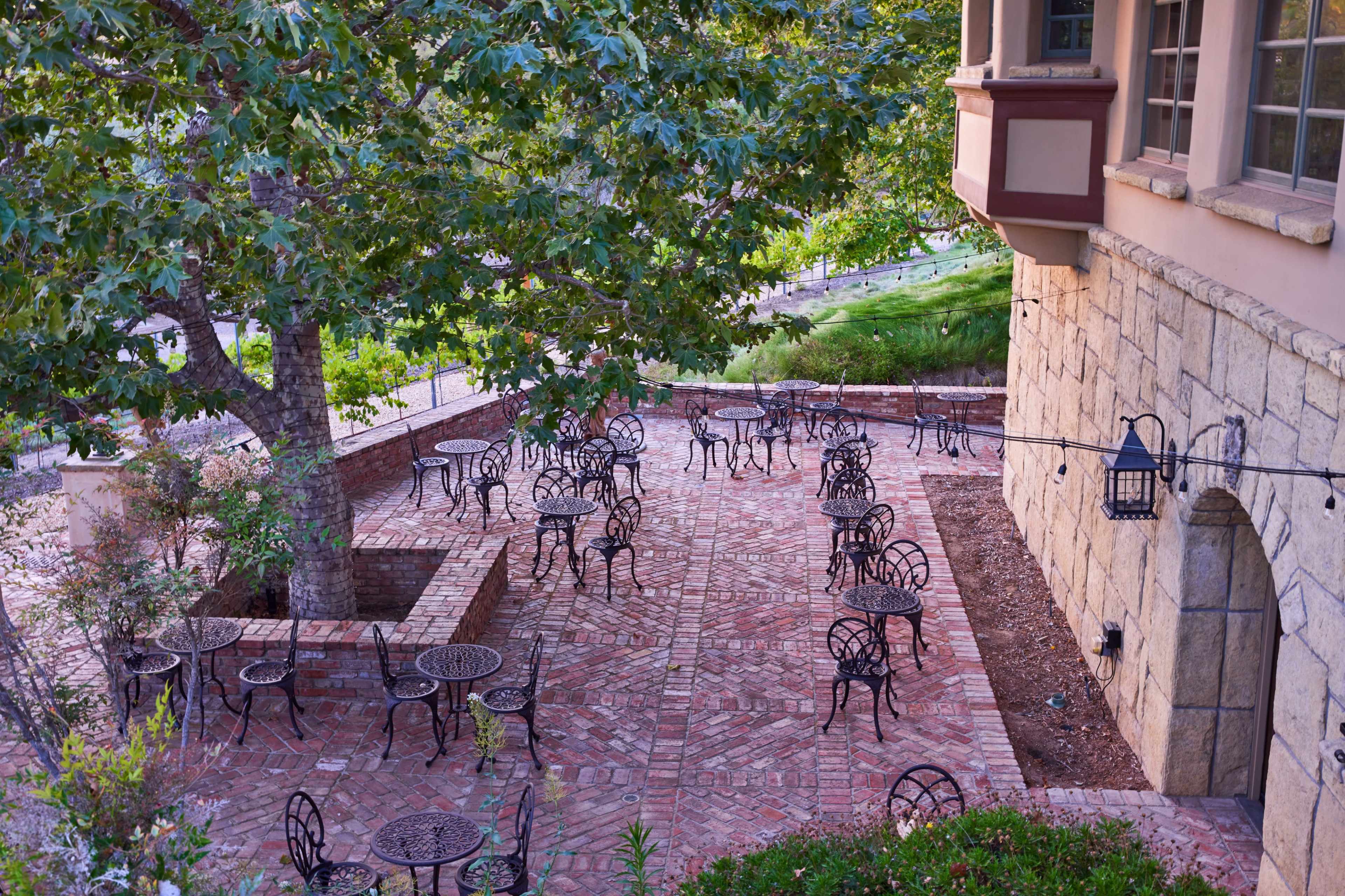 A brick patio with multiple round tables and chairs arranged under a large tree beside a stone building.