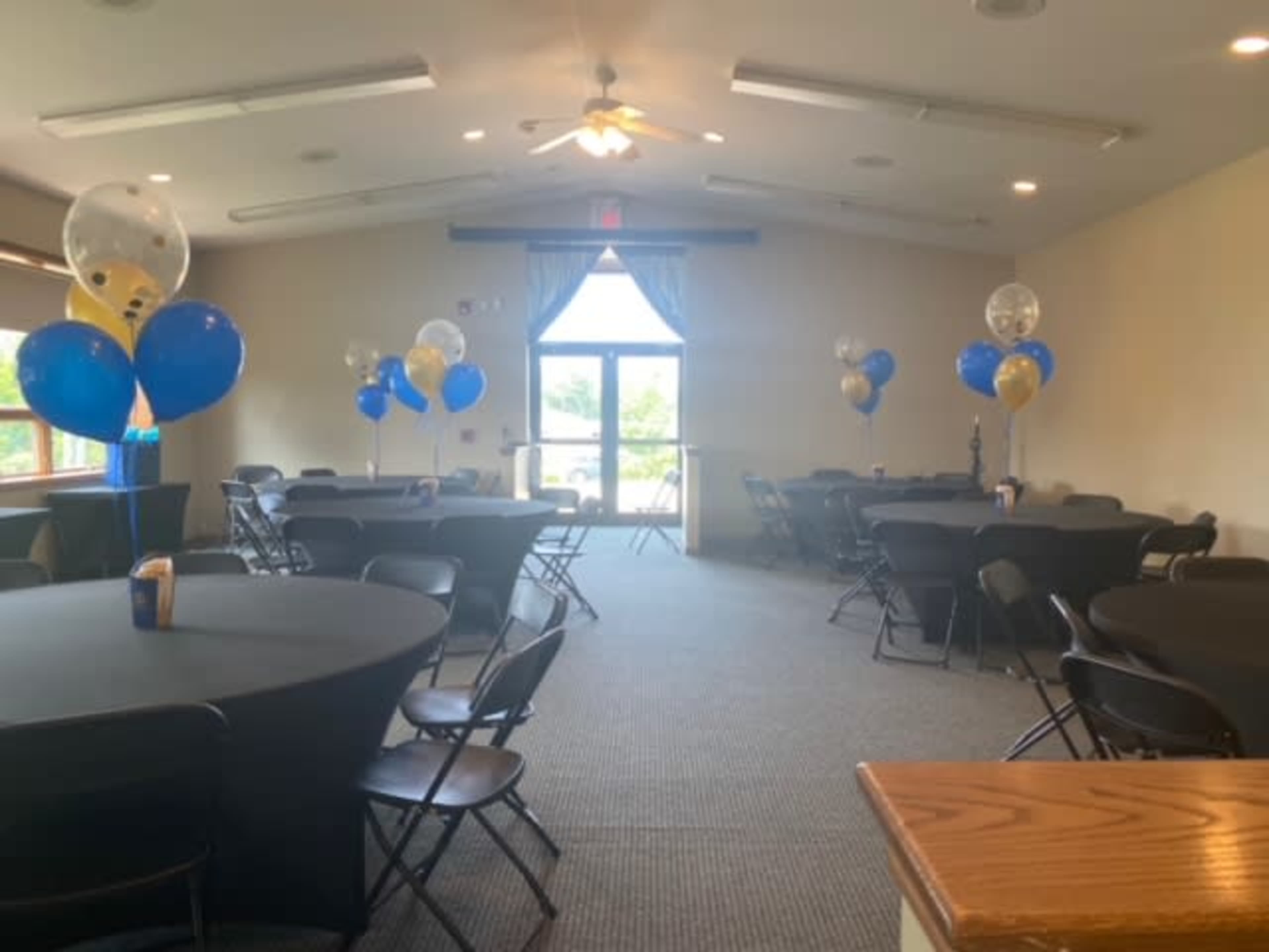 A spacious event room with several round tables covered in black tablecloths, decorated with balloons in blue and gold, and a large window at one end.