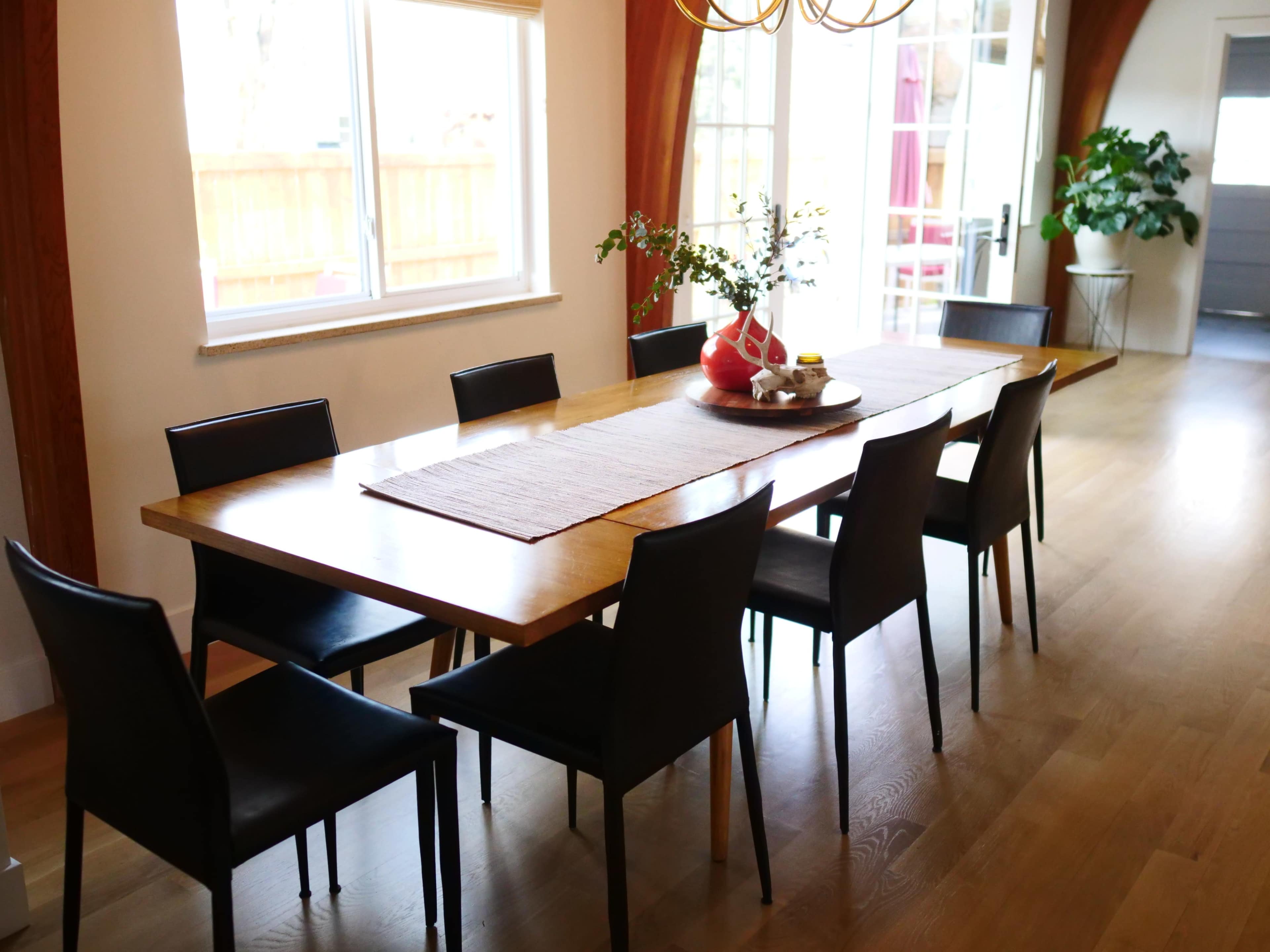 A long wooden dining table is surrounded by eight black chairs in a well-lit room with large windows.