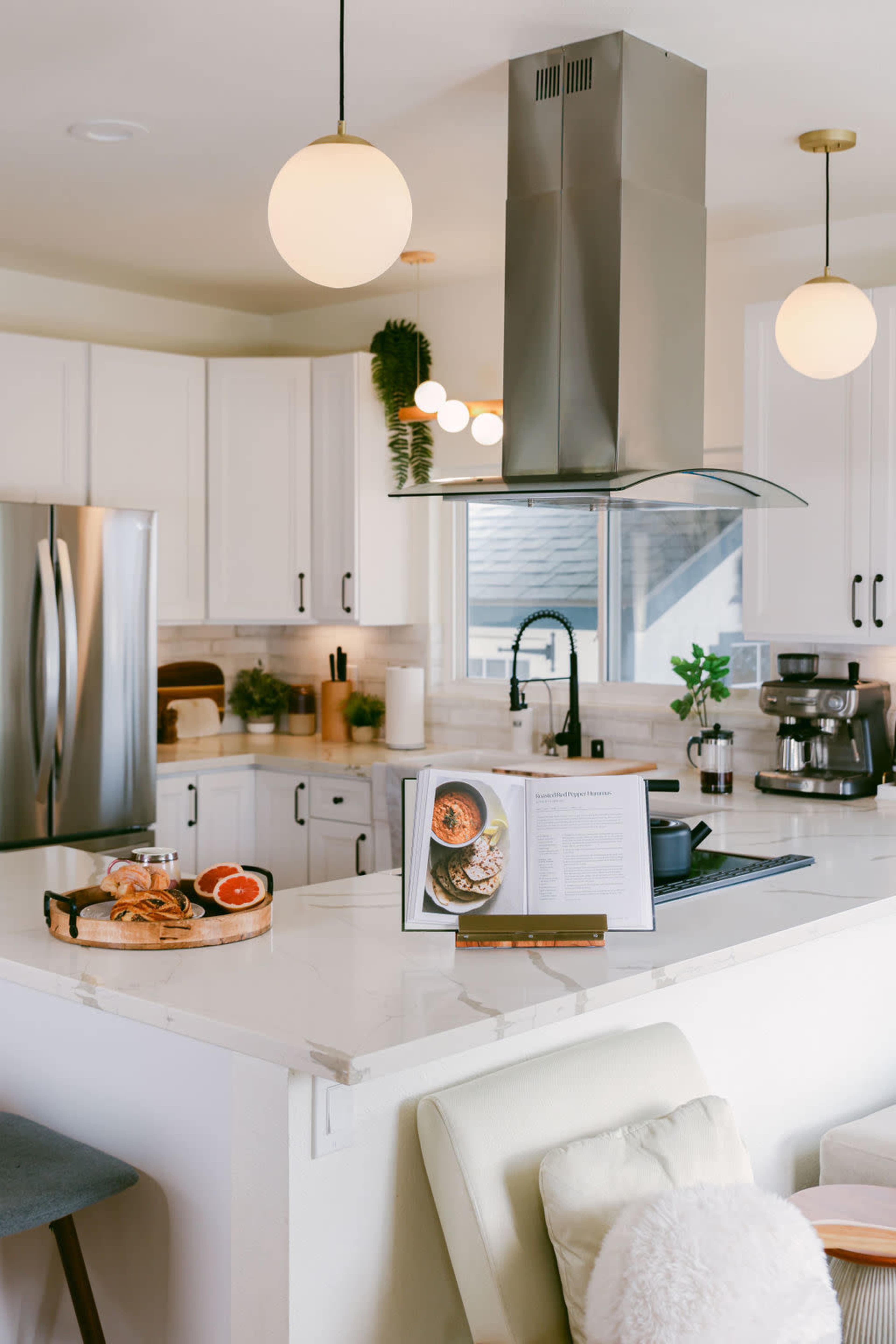 A modern kitchen features a central island with a cookbook and food items, stainless steel appliances, white cabinetry, and pendant lighting.