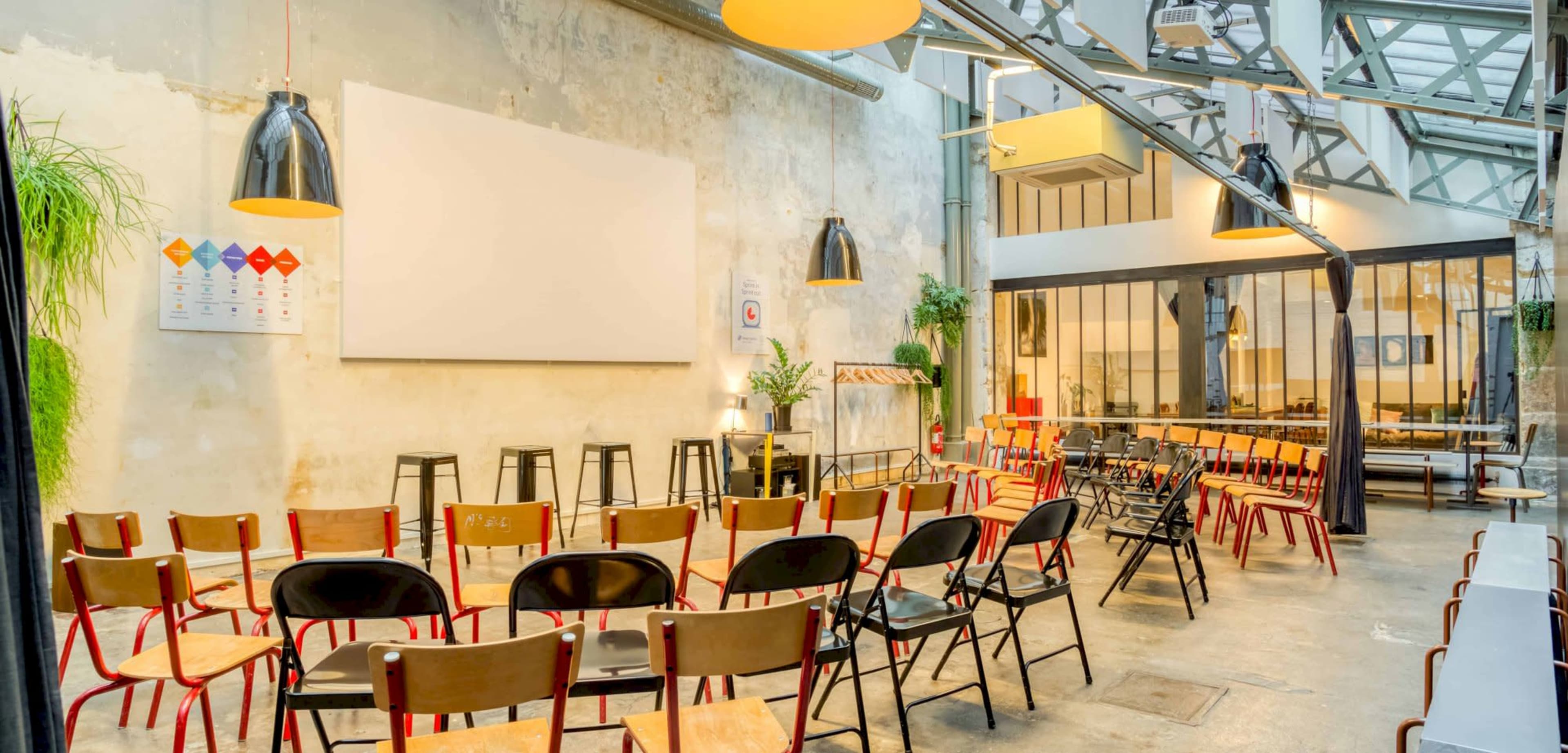 A modern event space features rows of wooden chairs arranged in front of a large screen against a textured wall, with plants and industrial lighting.