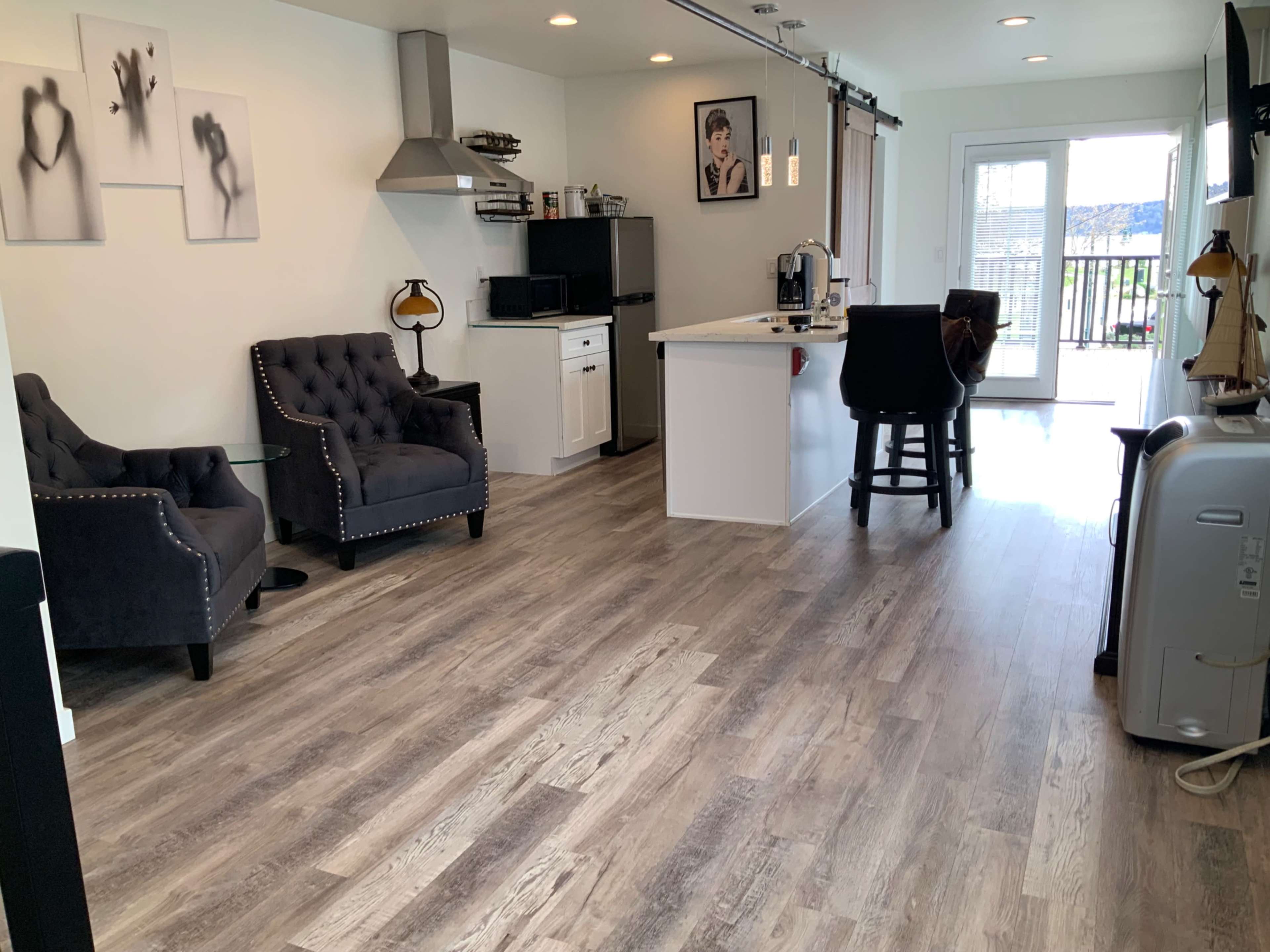A modern living space with two gray tufted armchairs, a small glass side table, a kitchen area with white cabinets, and a balcony door leading outside.