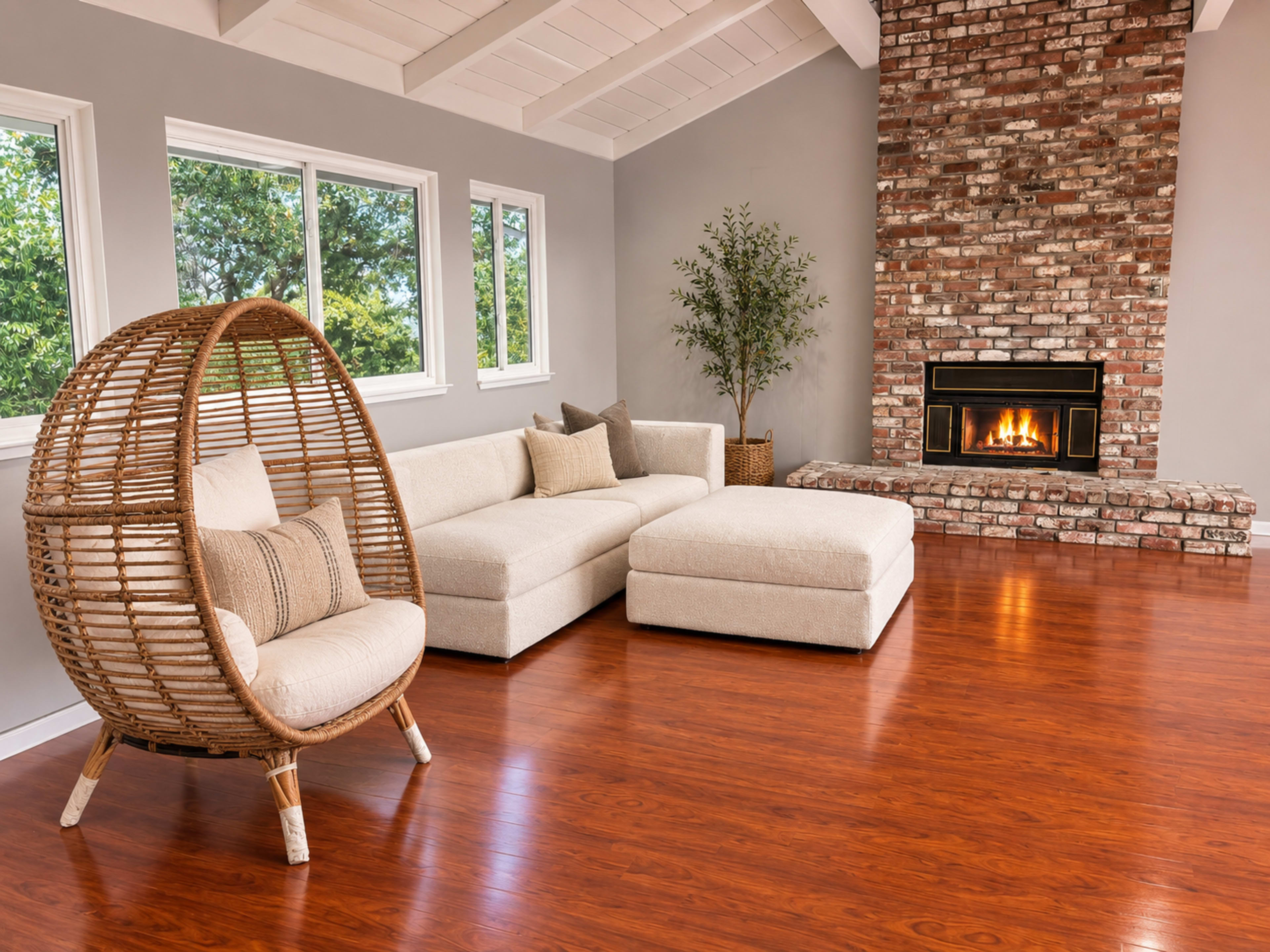 The image shows a modern living room featuring a light-colored sectional sofa, a round wicker chair, and a brick fireplace with a fire burning.