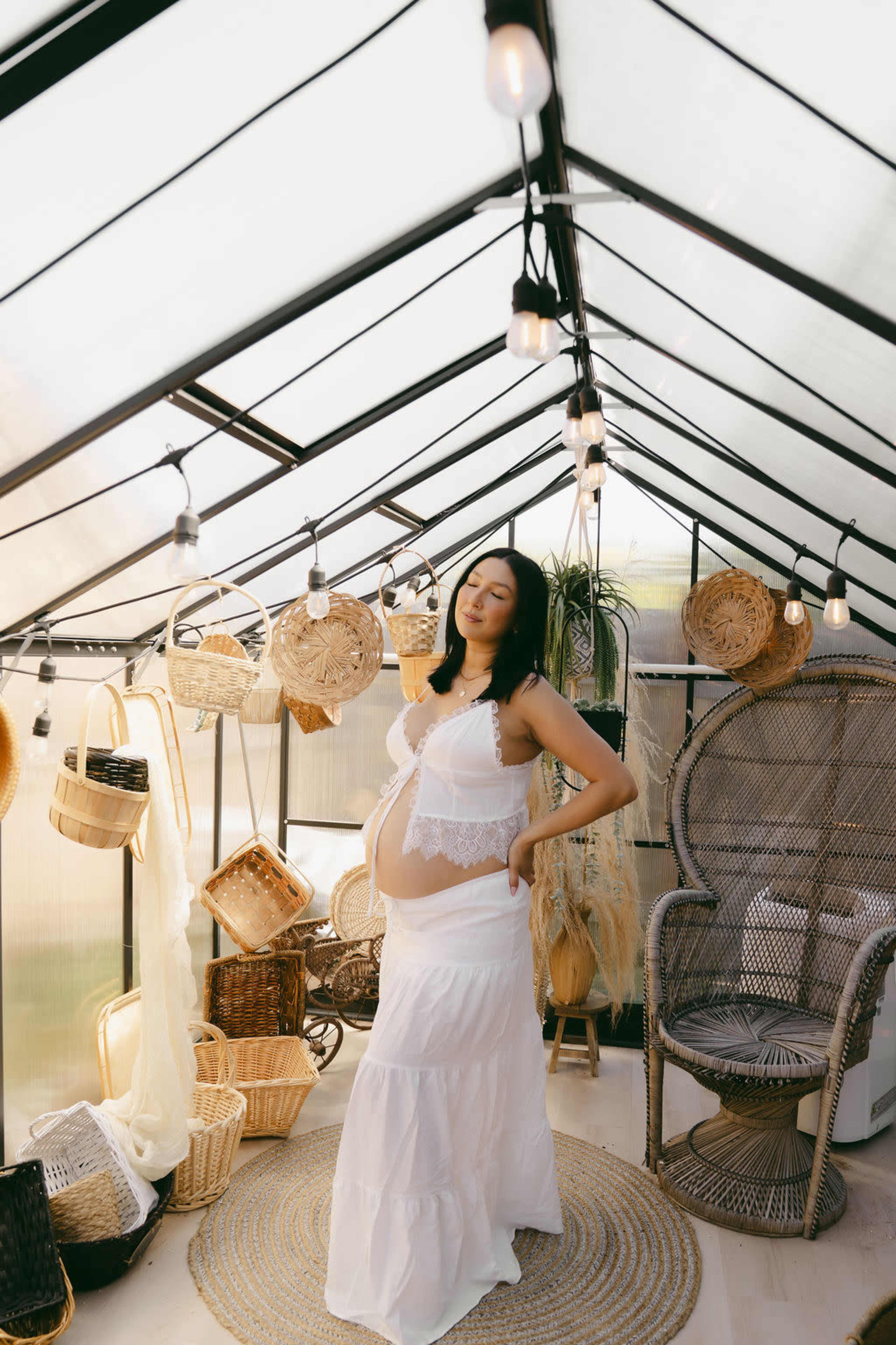 A woman stands in a greenhouse, showcasing her baby bump while wearing a white outfit amidst decorative baskets and plants.