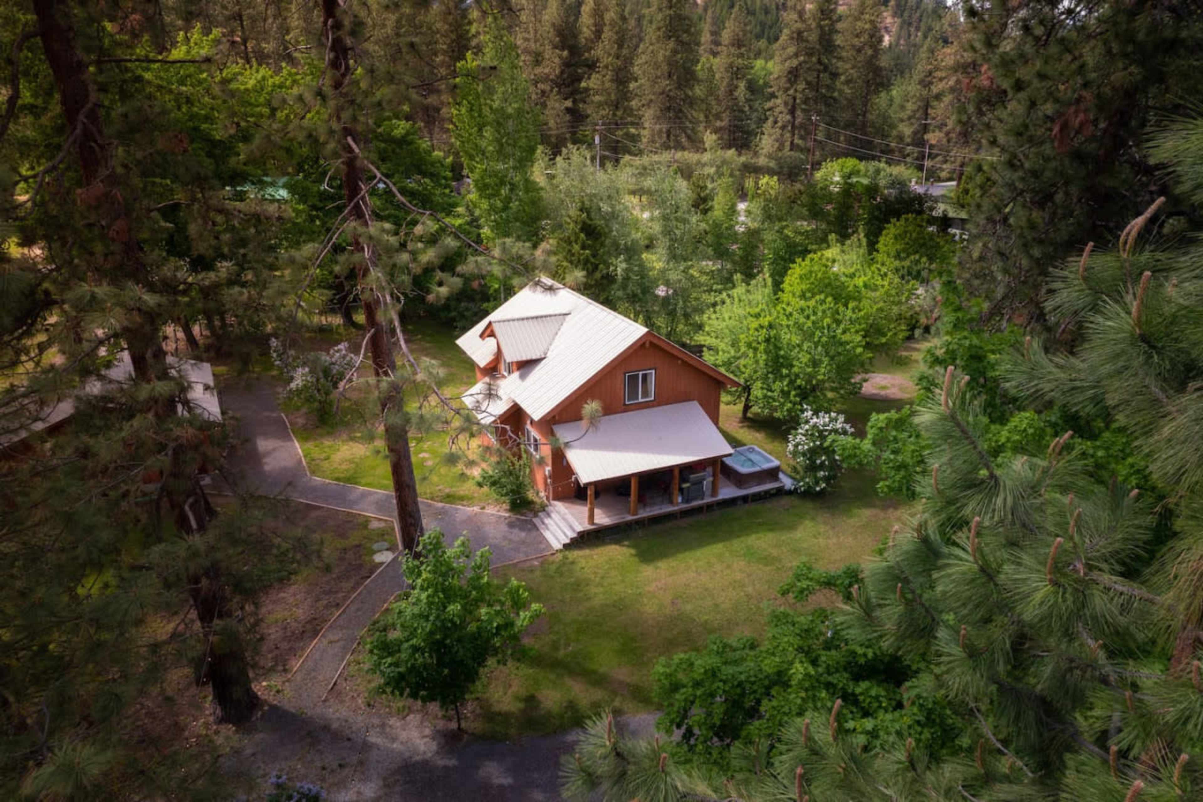 A two-story wooden house is surrounded by trees and greenery, with a pathway leading to it.