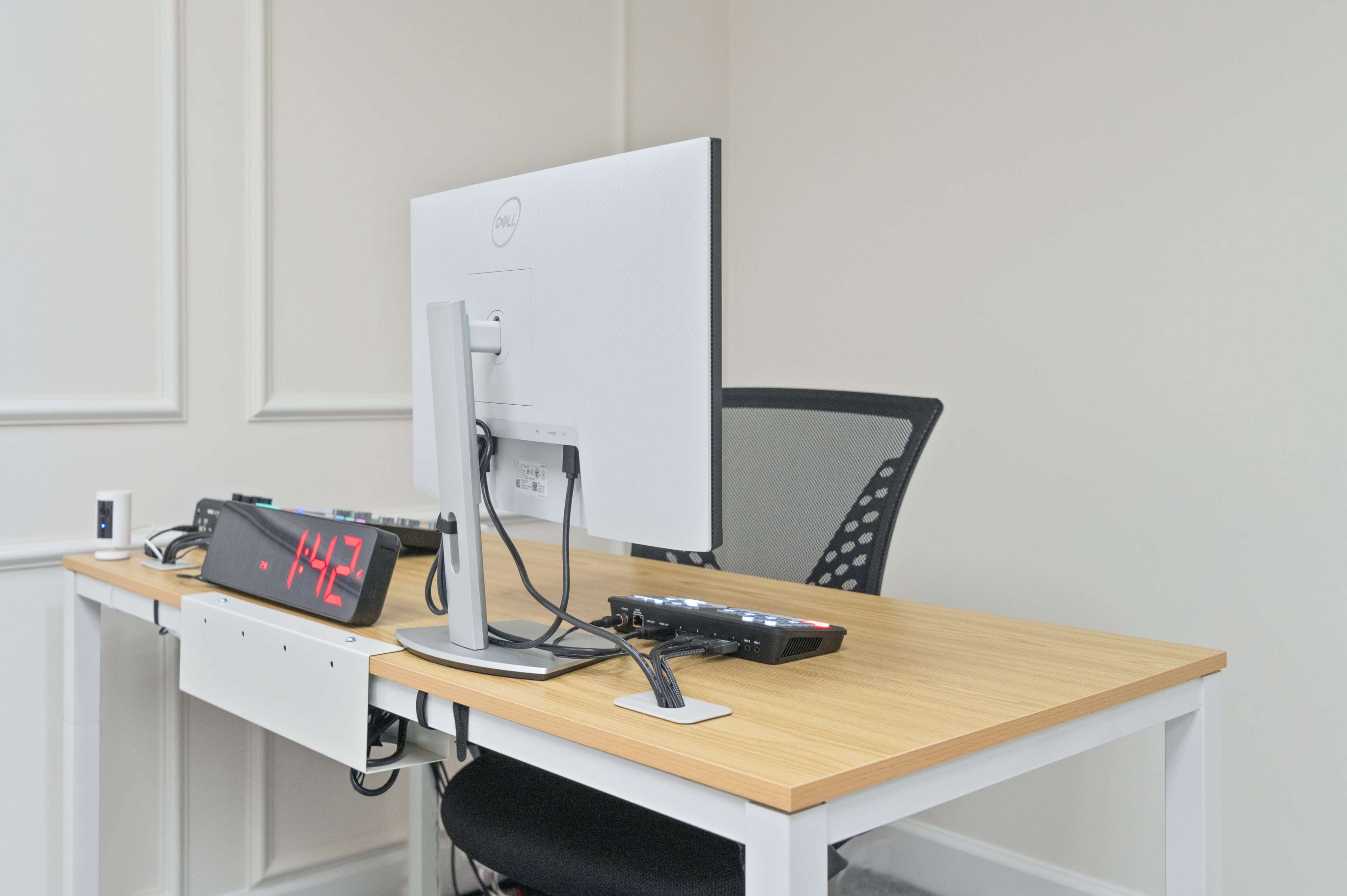 A desk setup features a computer monitor, a digital clock, and a chair in a minimalist office space.