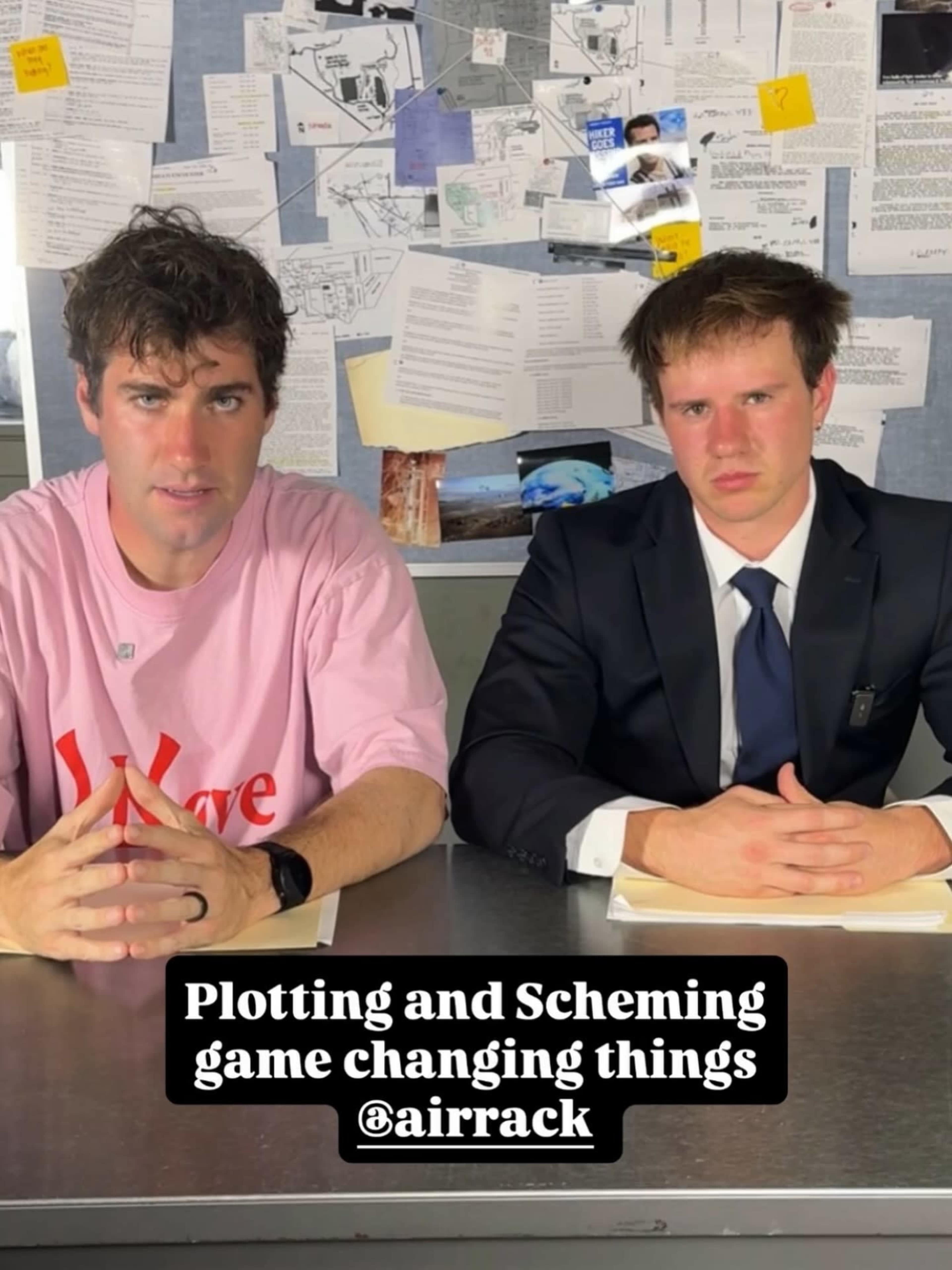 Two men are seated at a metal table, with one wearing a pink T-shirt and the other in a suit, as they appear to be discussing plans against a backdrop of papers and images pinned to a wall.