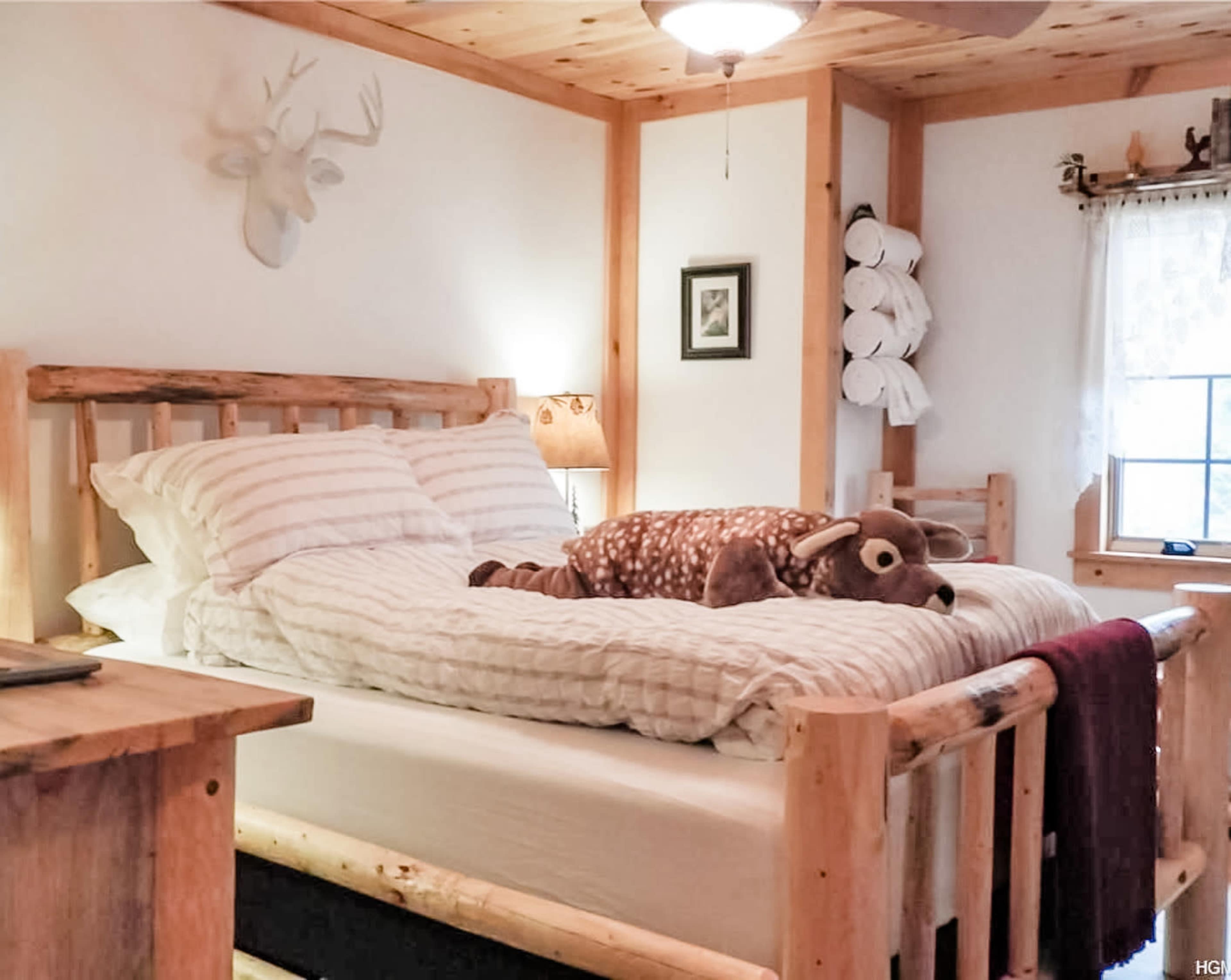 The image shows a rustic bedroom featuring a log bed with a plush stuffed animal lying on top of the bedding, surrounded by wooden decor and a window.