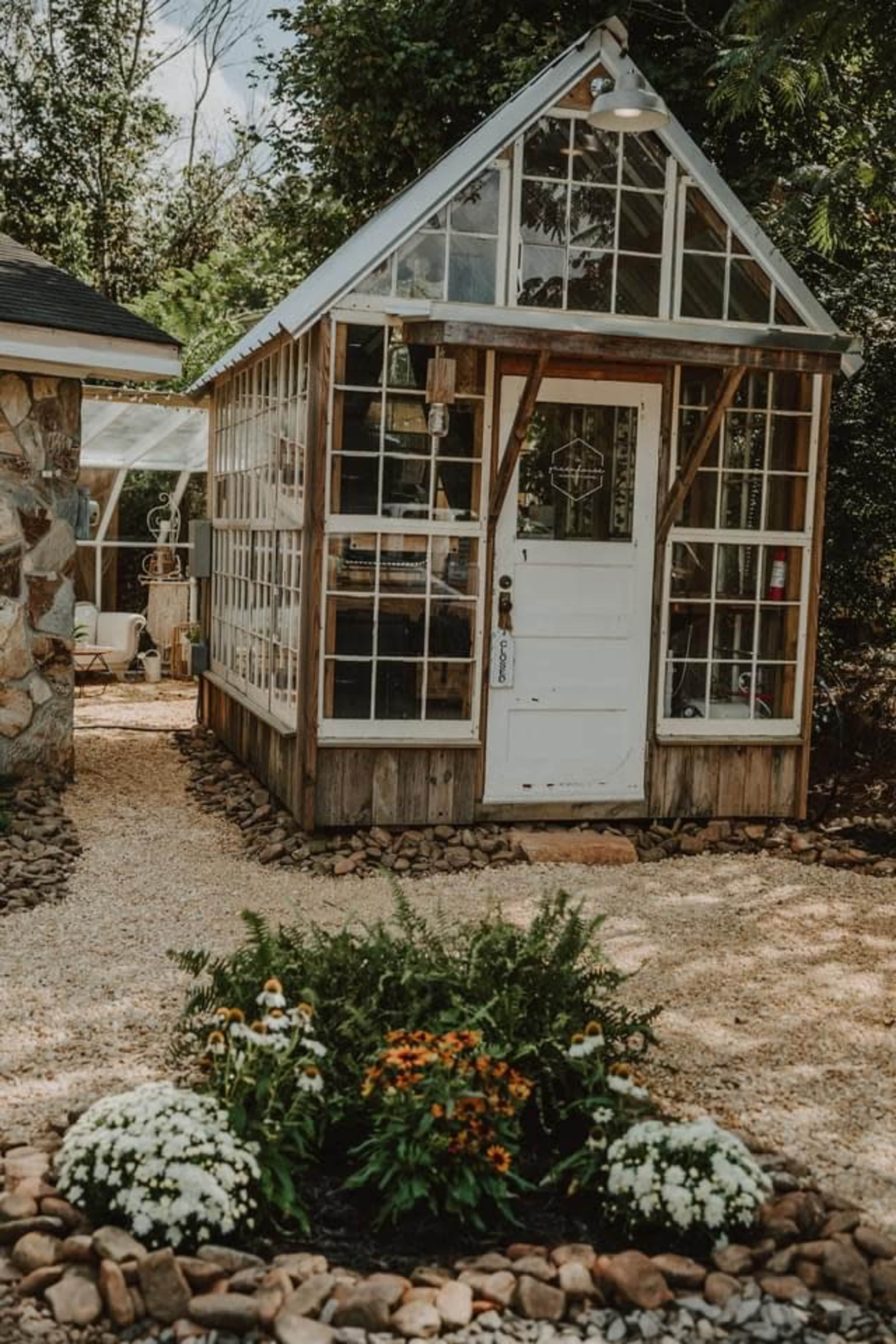A wooden greenhouse with glass panels stands in a gravel yard, surrounded by flower beds and greenery.