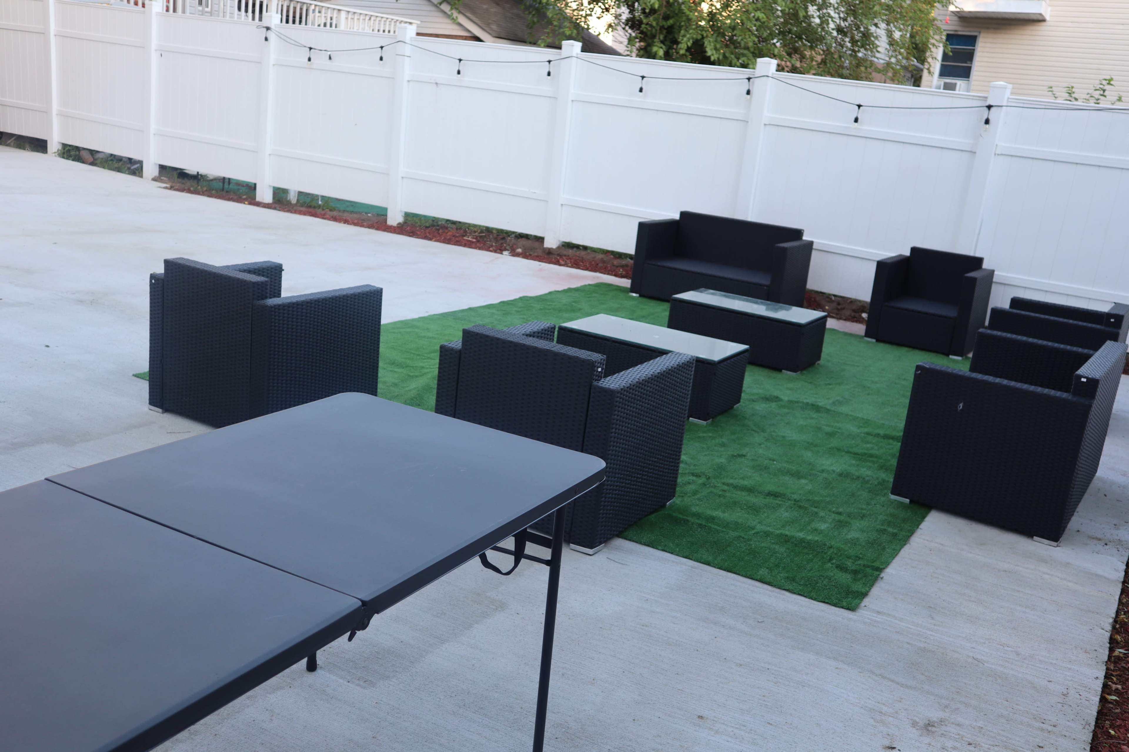 A paved outdoor space features a green artificial grass area surrounded by black wicker furniture, including sofas and armchairs.