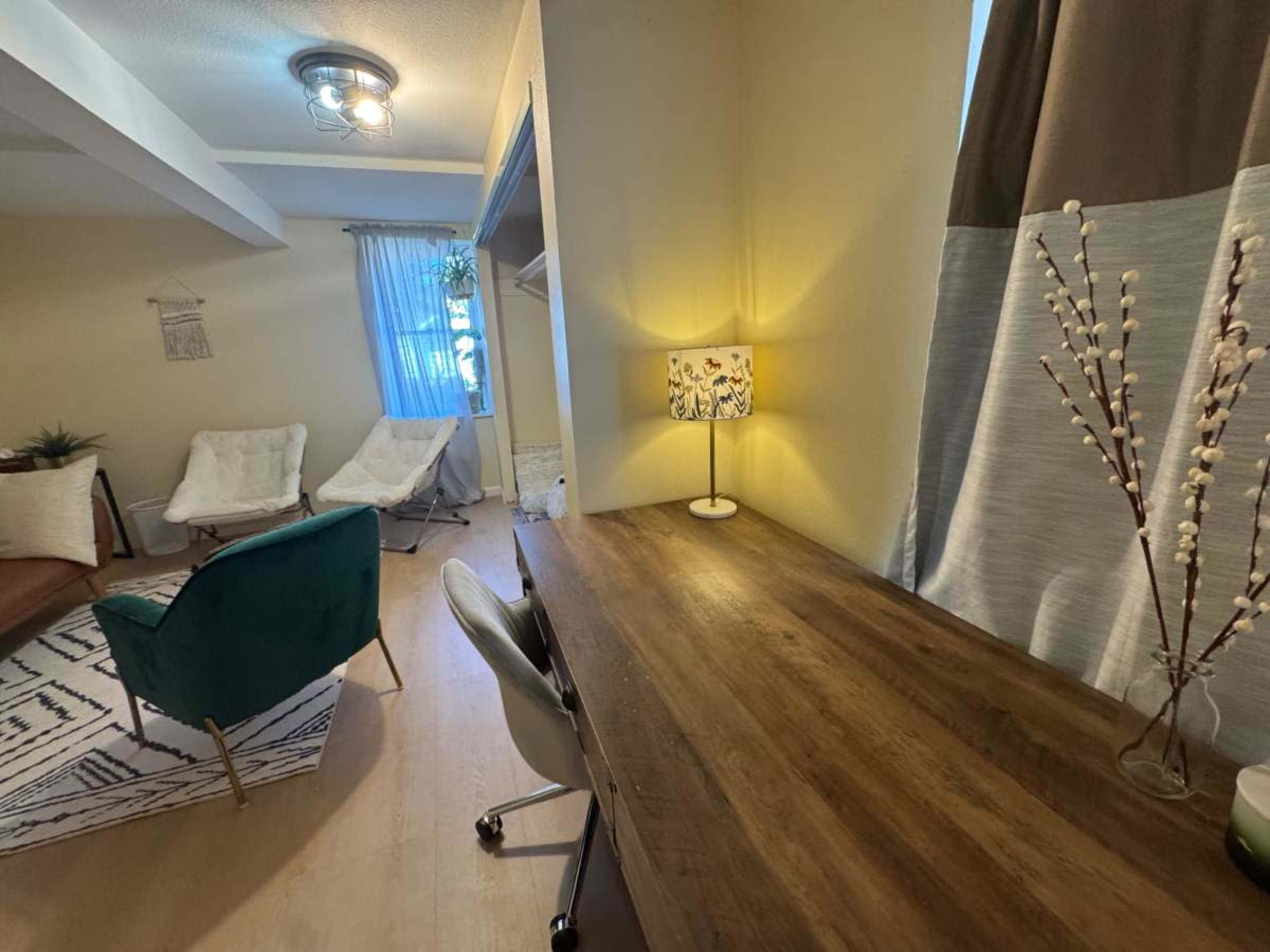 The image shows a cozy living space with a wooden desk, a decorative lamp, and a couple of modern chairs, all illuminated by natural light from a window.