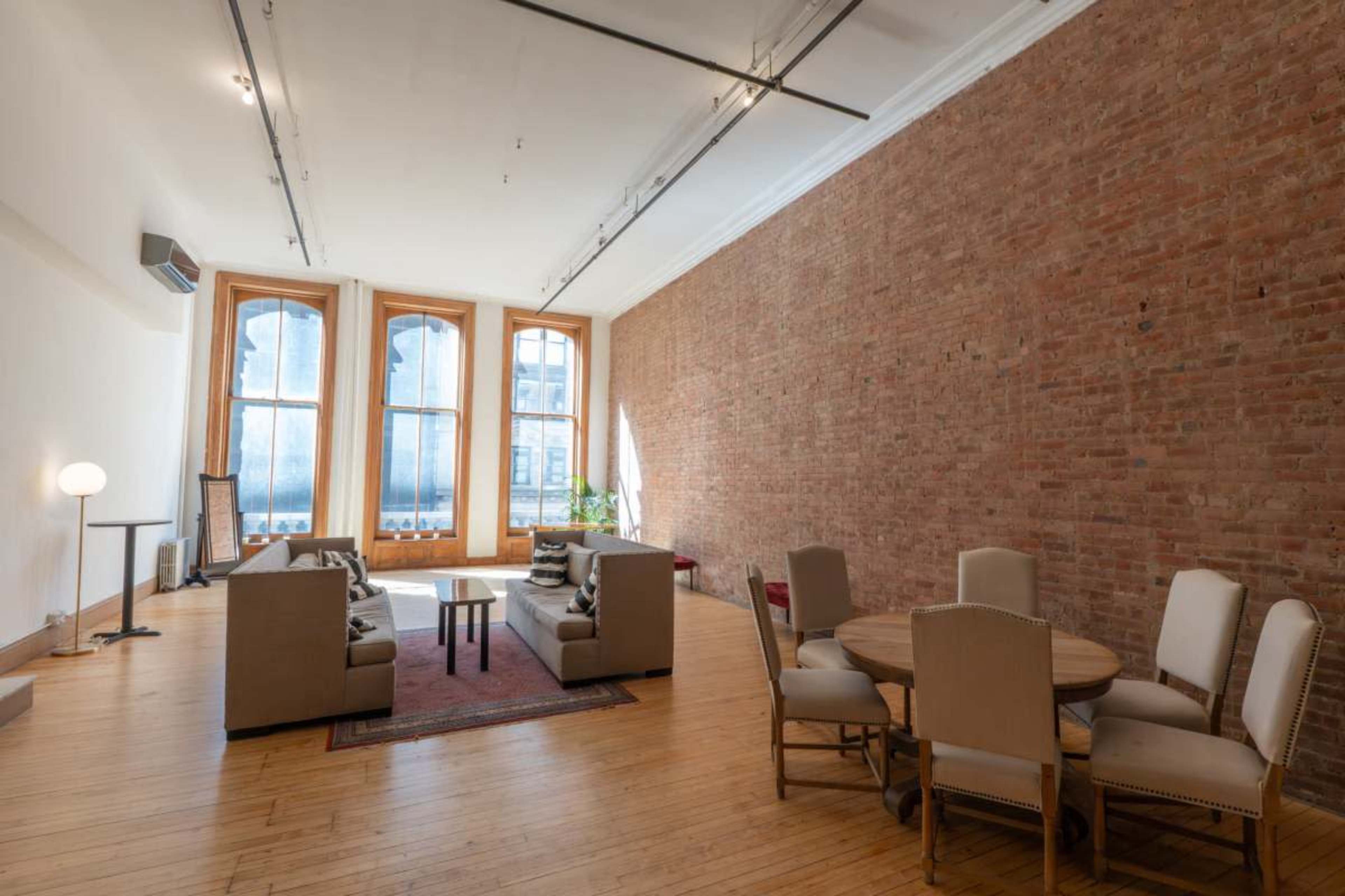 The image depicts a spacious room with exposed brick walls, large windows, a seating area with sofas, and a dining table with chairs.