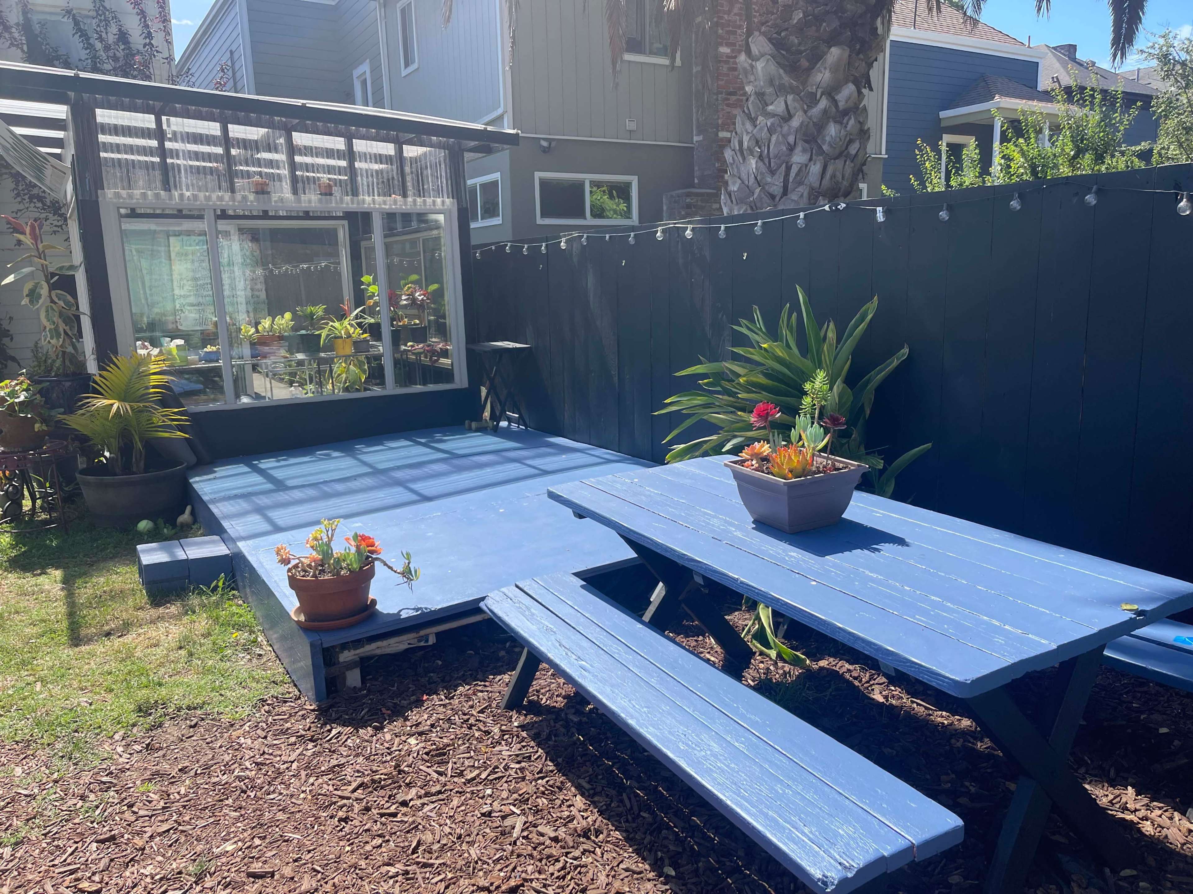 A blue wooden deck with a picnic table and potted plants is set in a backyard next to a greenhouse and surrounded by a black fence.