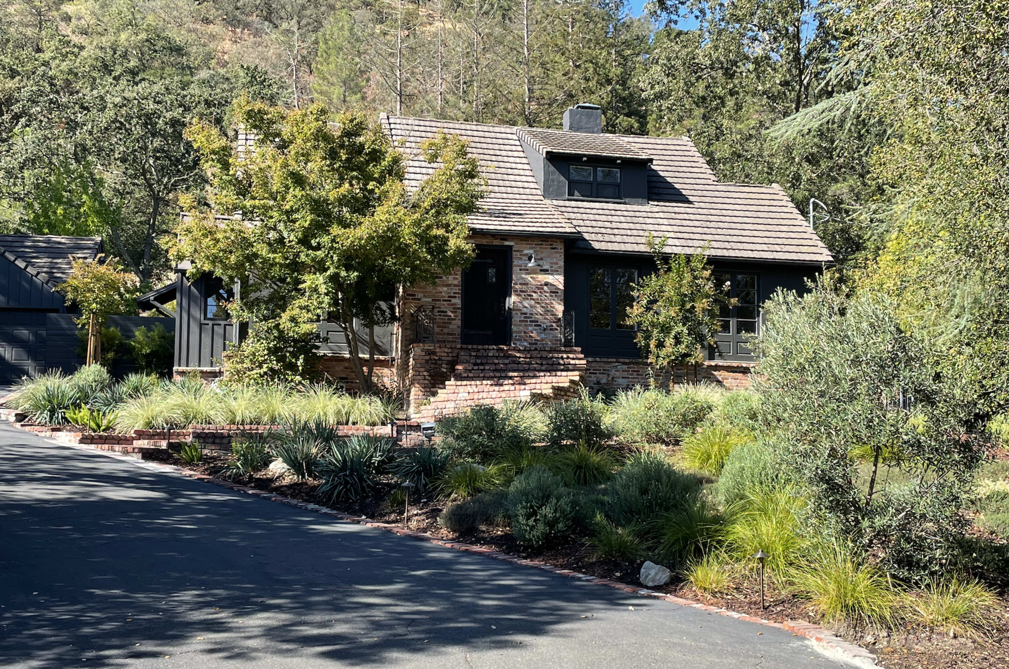 A two-story, dark-hued house with a brick facade and a sloped roof is set among greenery on a winding driveway.