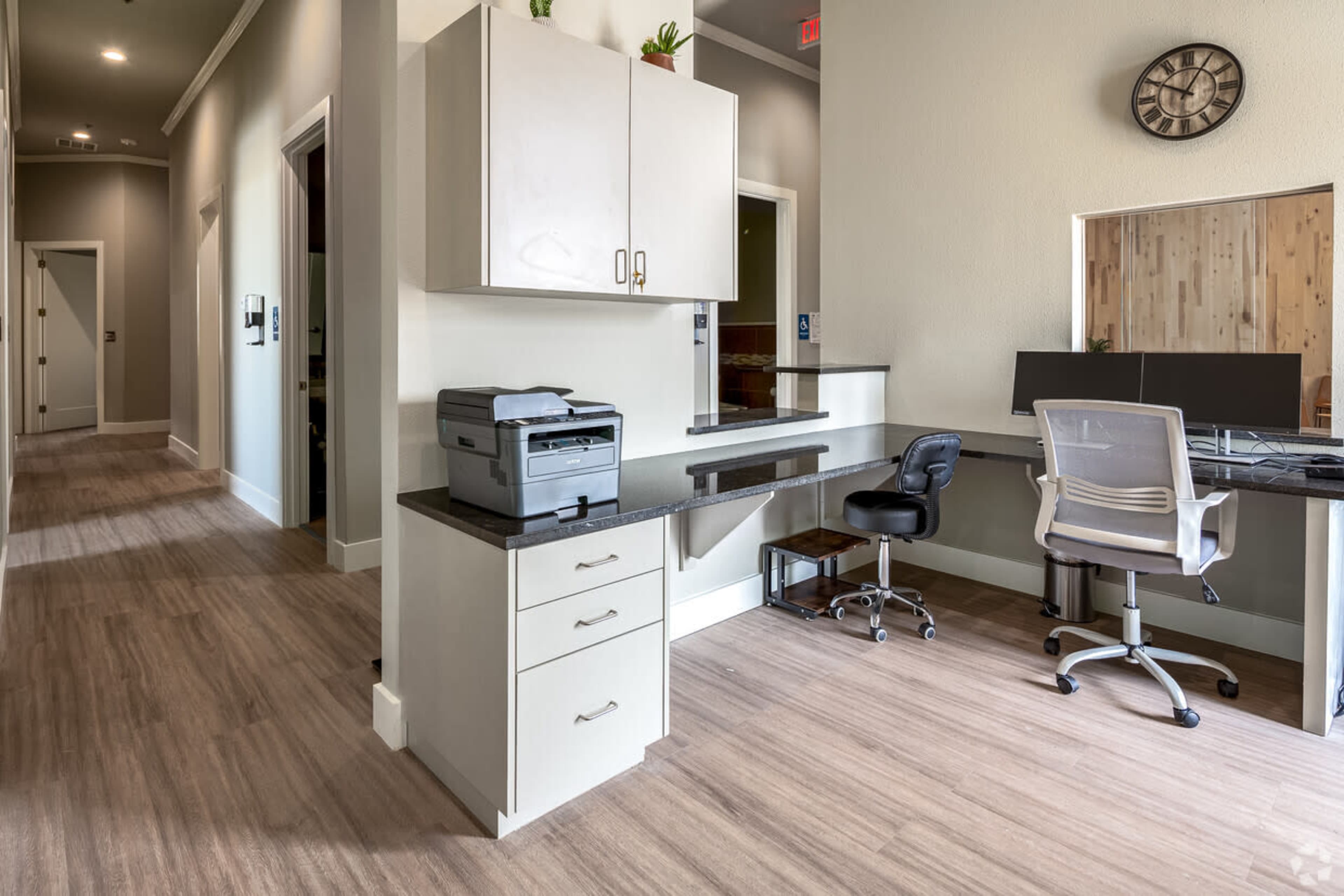 A modern office space featuring a printer on a countertop, a desk with a computer setup, and light wood flooring.