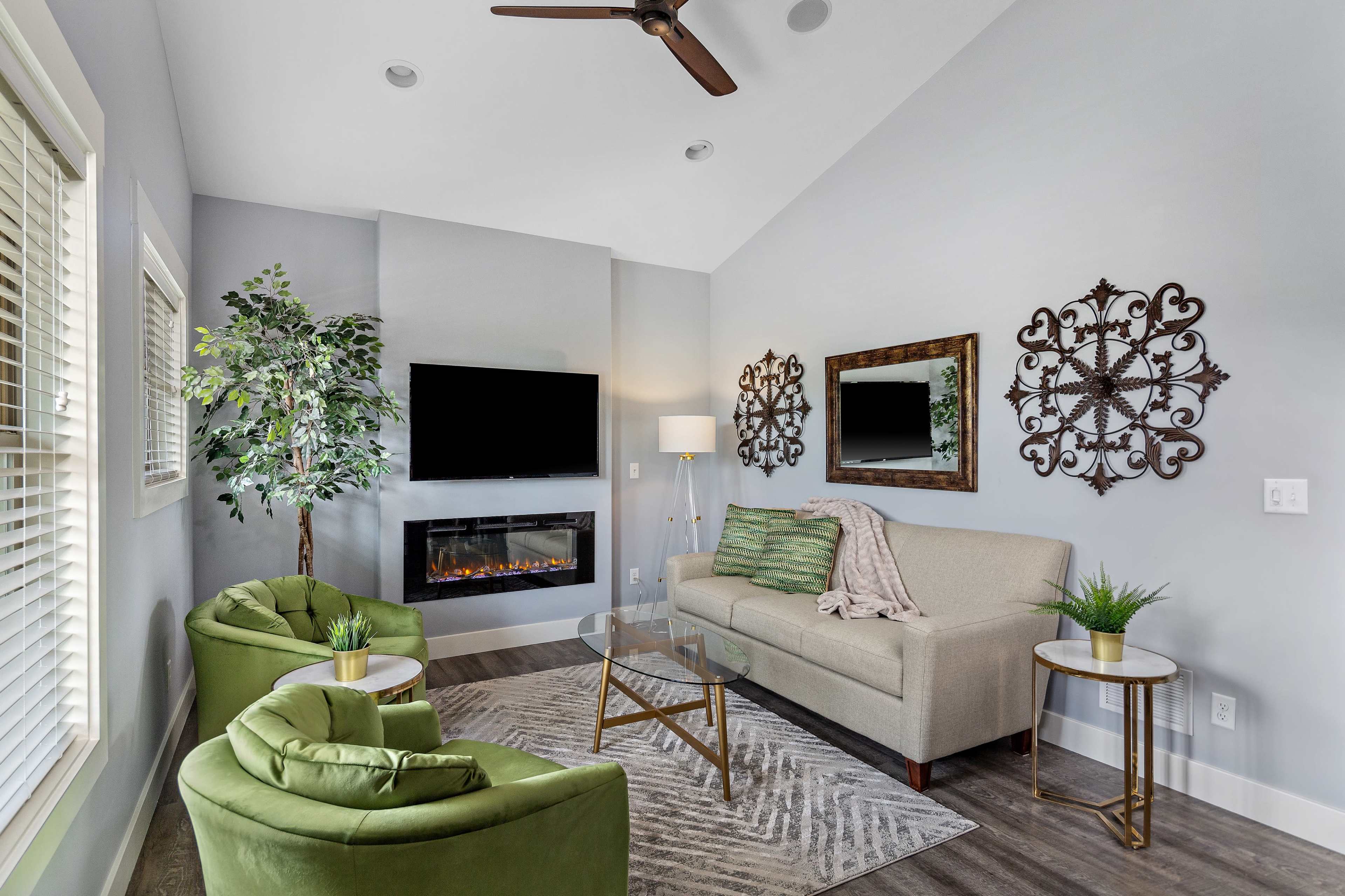 A modern living room featuring a green sofa and armchairs, a wall-mounted television, an electric fireplace, and decorative mirrors on the walls.