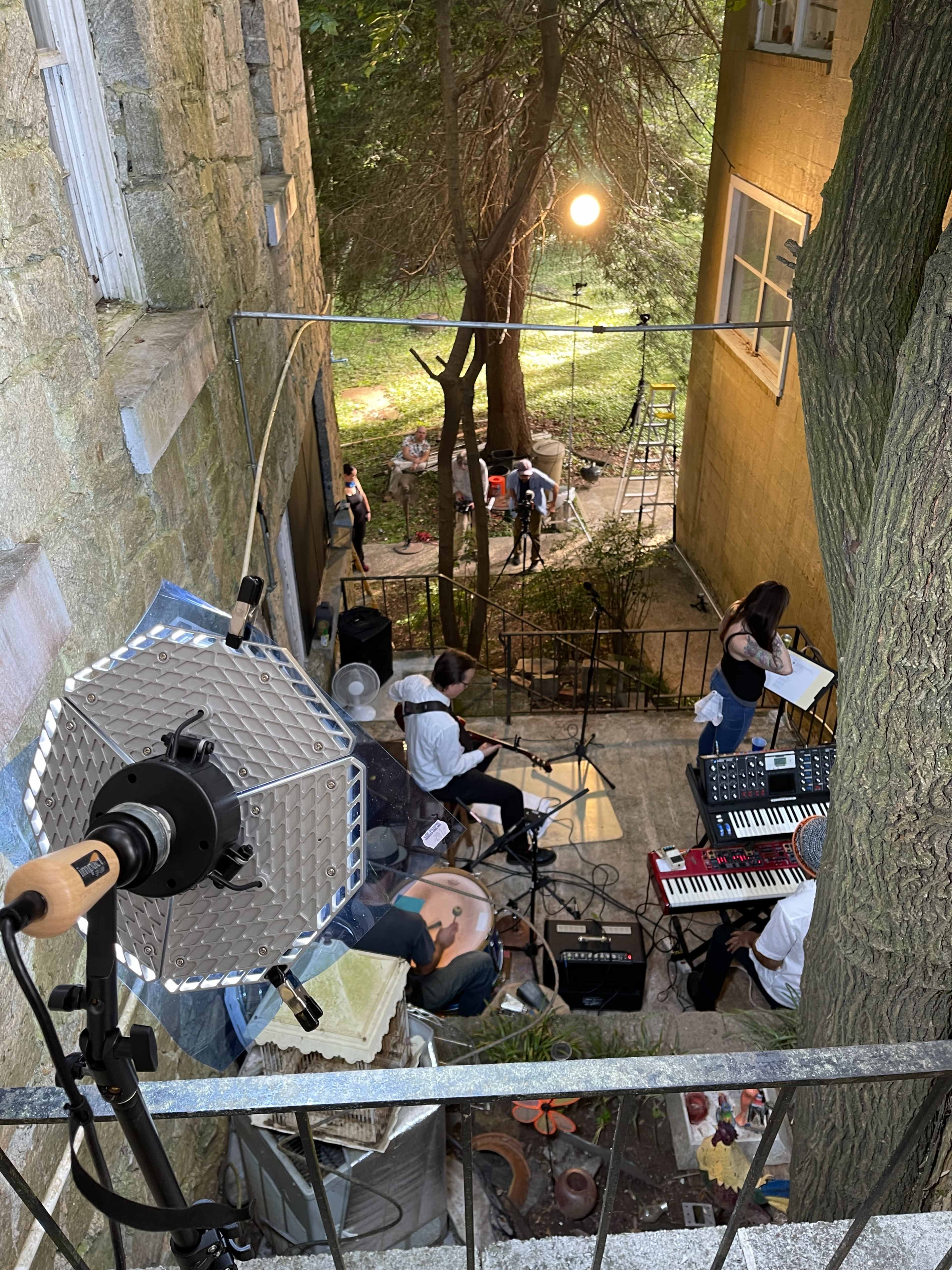 A group of musicians performs in a courtyard surrounded by trees and a building, with a spotlight illuminating the area.