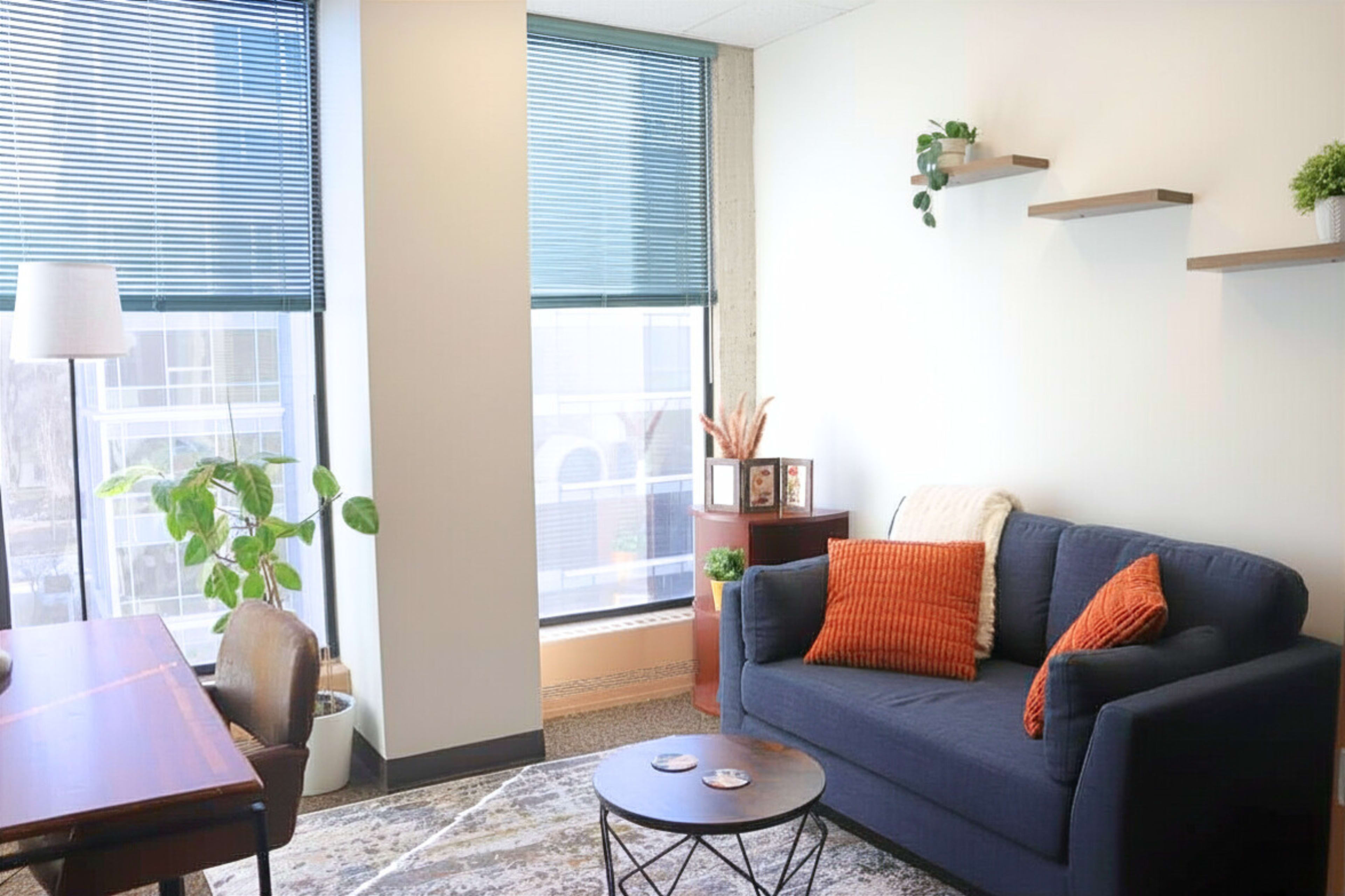 The image shows a modern office space featuring a dark blue sofa with orange pillows, a wooden desk with a chair, and a large window with blinds, complemented by small indoor plants and shelves on the wall.