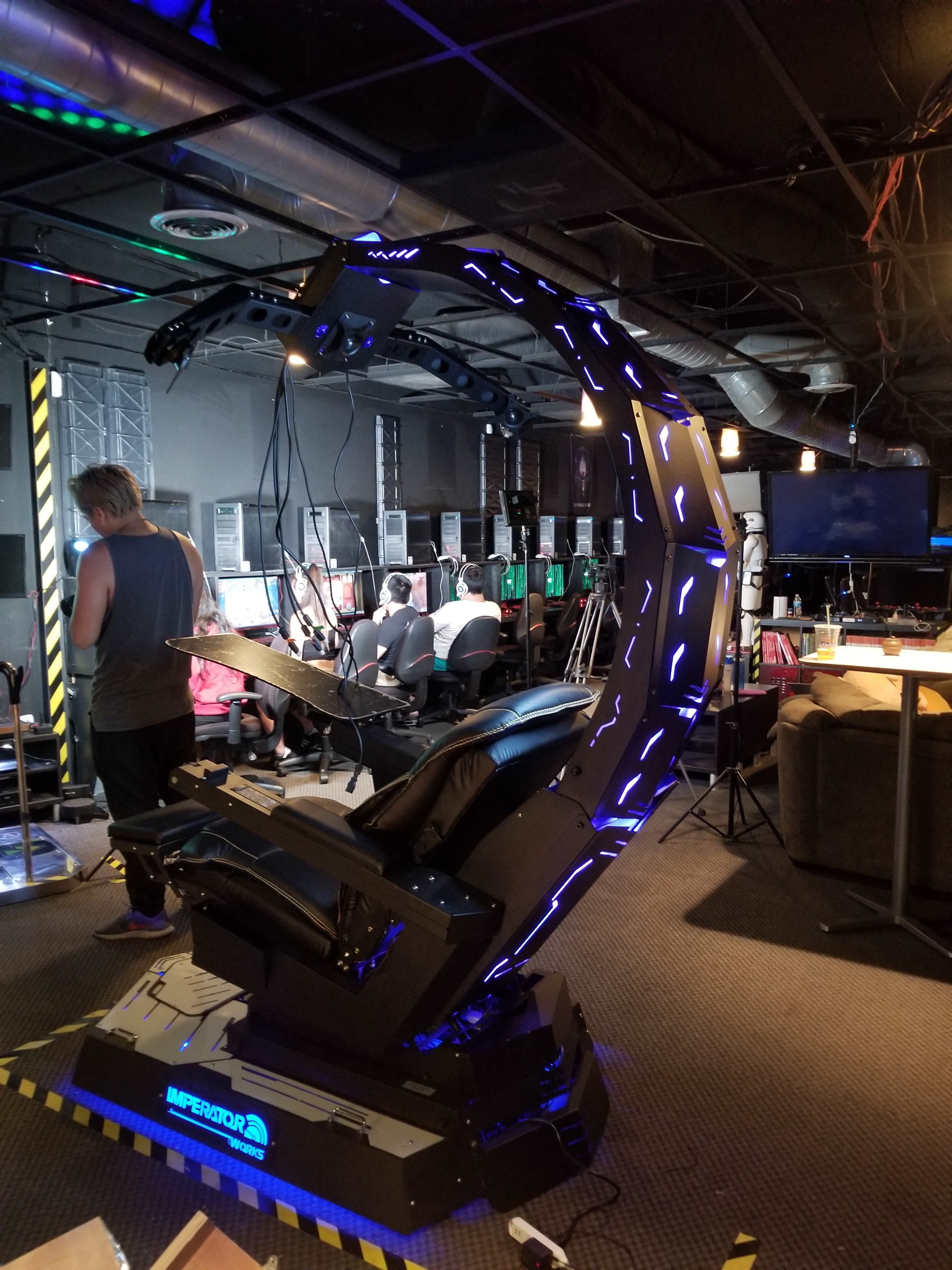 A futuristic gaming chair with neon lighting is positioned in a room filled with computers and people engaged in gameplay.
